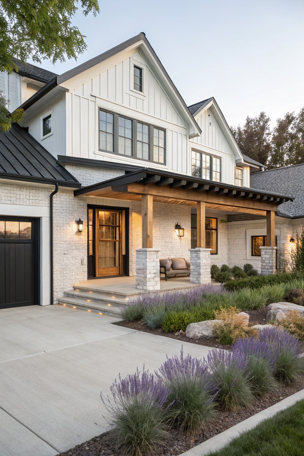 White board-and-batten sided house with black metal roof sections, covered front porch supported by stone columns and wood beams, attached dark garage door, concrete driveway, and landscaped beds with lavender plants and rocks.