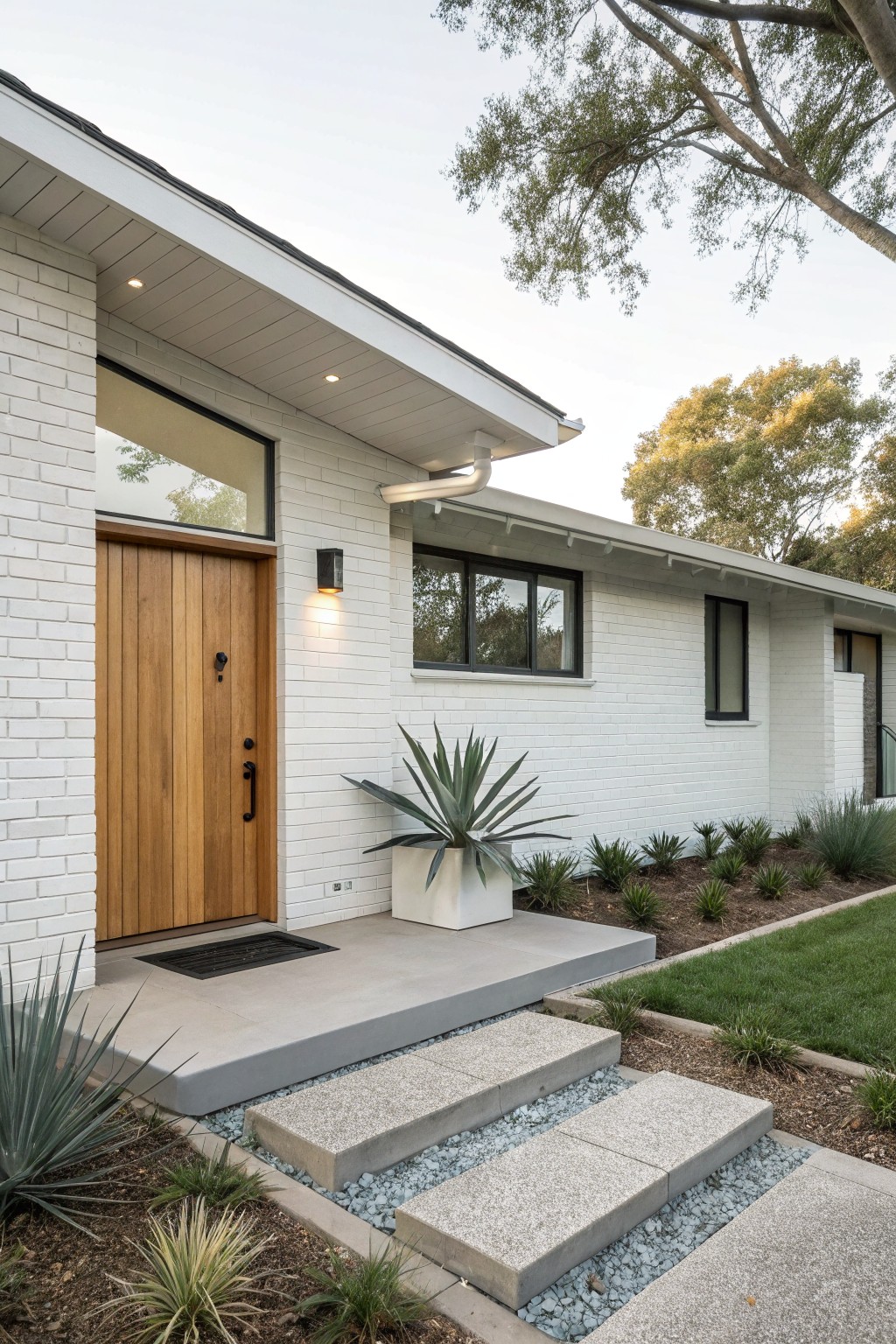 Ranch-style house exterior featuring white painted brick walls, a vertical plank wooden front door with black hardware, a large fixed window beside the door, concrete entry steps with pebble accents, and agave plants in white pots near the base.