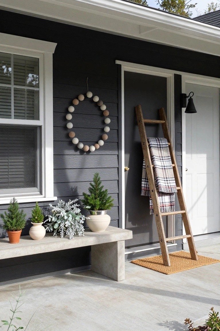 Dark gray house front porch with white-trimmed door and window, beige beaded wreath hanging on door, leaning wooden ladder draped with plaid blanket, small potted Christmas trees and plants on concrete bench, lantern light fixture, and seagrass doormat on concrete landing.