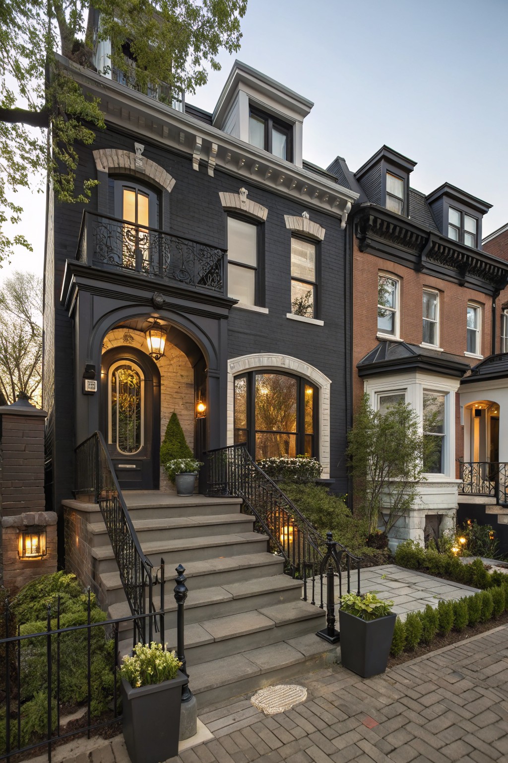 Dark painted brick rowhouse exterior with arched entry door, wrought iron balcony railings and stairs, stone steps, potted plants, and landscaping along a brick pathway at dusk.