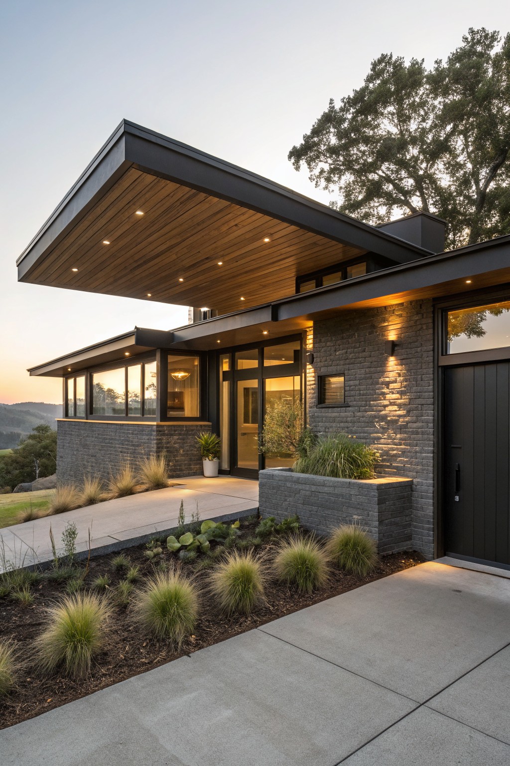 Modern house exterior with dark brick walls, cantilevered black metal roof with wooden ceiling, large glass entry doors and windows, brick planters with grasses, concrete driveway and path at dusk.