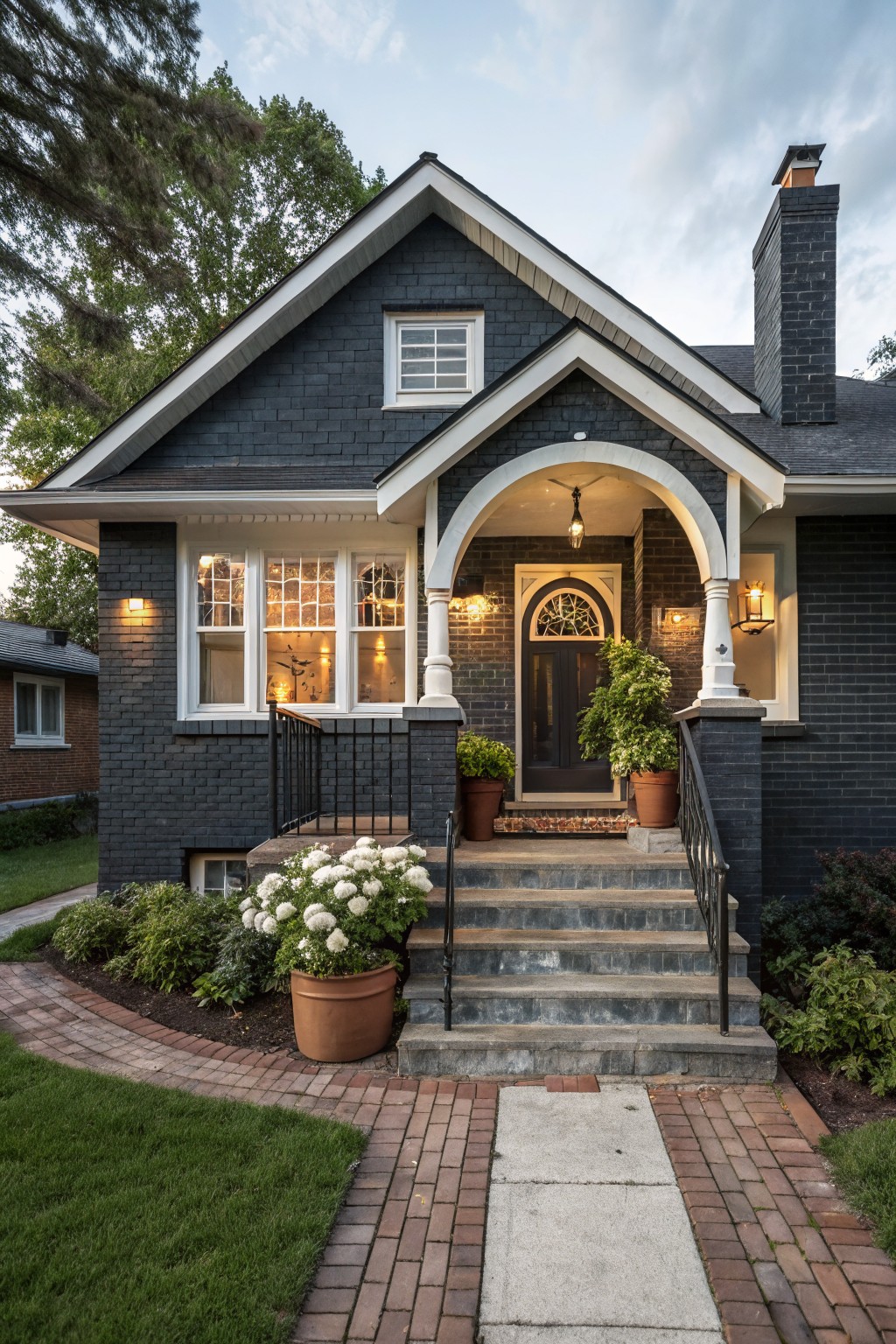 Dark gray brick Craftsman-style bungalow house with white trim, arched front porch entry, lit lanterns, potted plants on concrete steps, brick pathway, and surrounding landscaping at dusk.