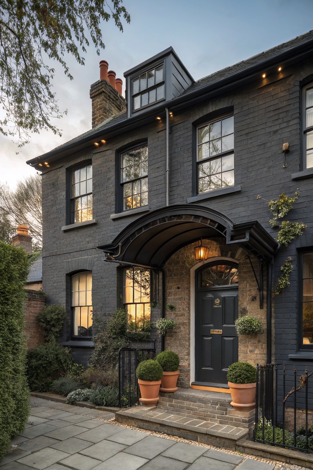 A two-story dark brick house exterior with black-framed sash windows, a curved black metal canopy over the dark wood front door, brick chimneys, exterior lights, potted plants on steps, hedges, and a paved pathway in evening light.