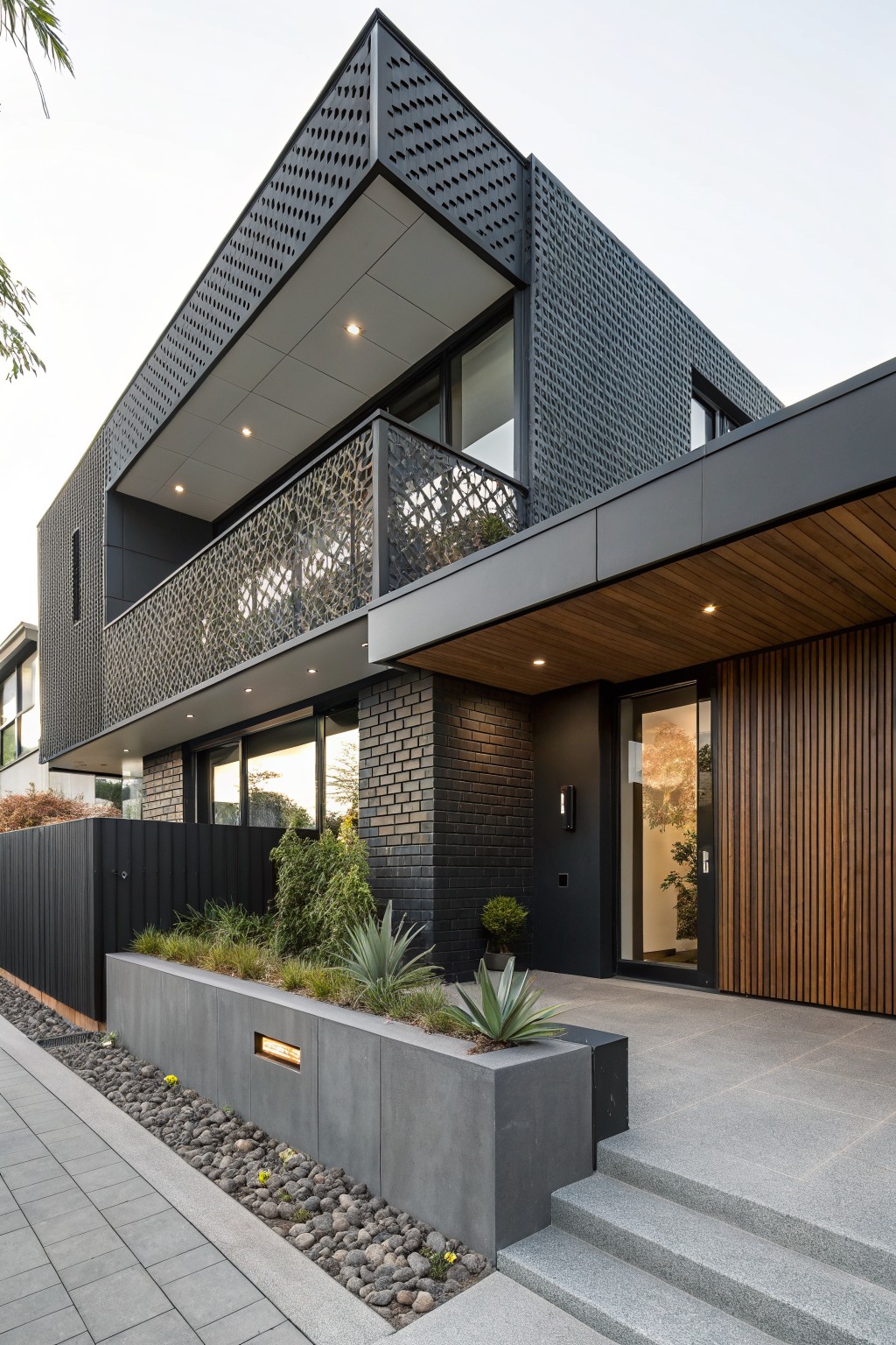 Modern two-story house exterior with dark brick walls, black perforated metal screens on upper facade and balcony, wood-clad entryway with glass door, concrete planter wall with agave plants, and gray stone steps.