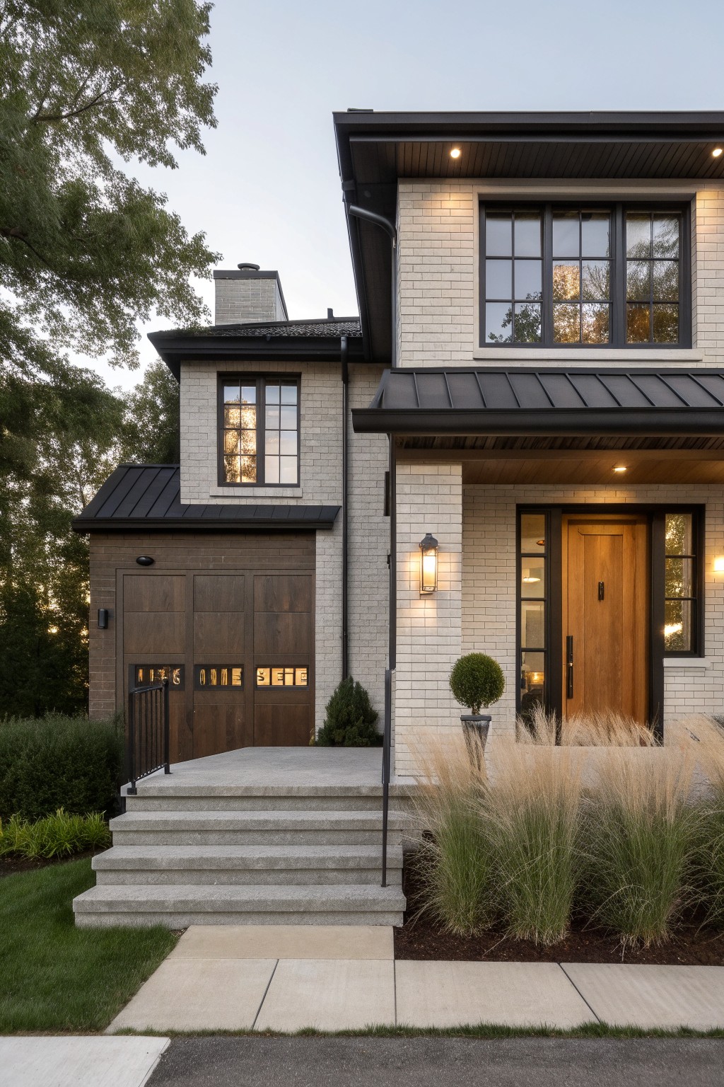Two-story brick house exterior with black metal roof, dark wood garage door, concrete steps to wooden front door with sidelights, lantern lights, ornamental grasses, and shrubs in front yard at dusk.