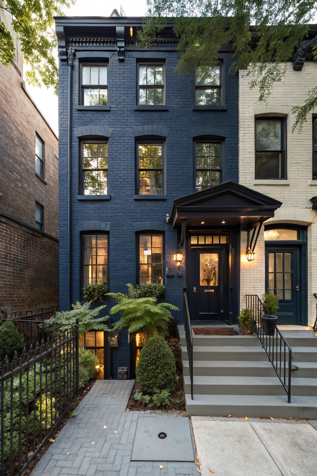Dark navy painted brick three-story townhouse with black window frames, covered entry porch, iron fence, potted plants, and steps, adjacent to lighter brick rowhouses under trees.