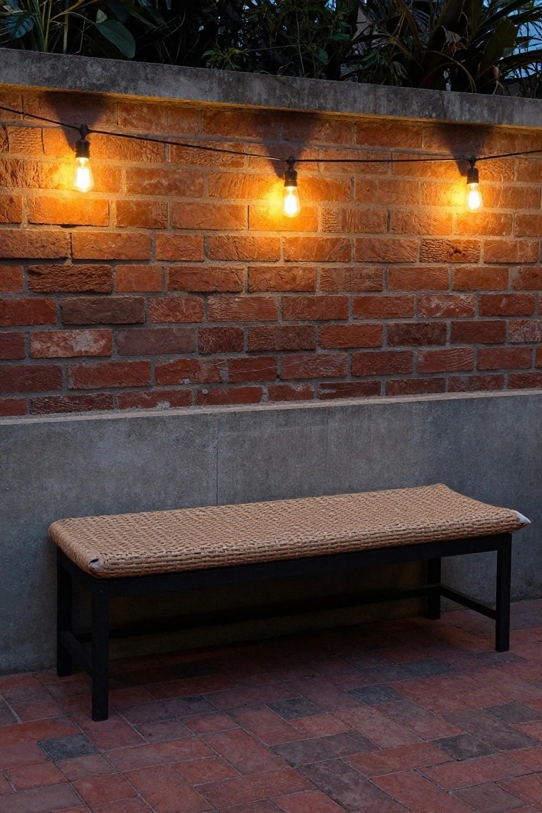 Brick wall with concrete base and greenery above, string lights with warm bulbs hanging along it, rattan bench on brick paver patio below