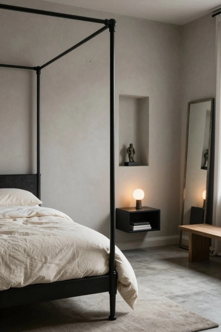 A minimalist bedroom with a tall black metal pipe-frame canopy bed draped in white linens, a wall niche holding a bronze male figure sculpture, a black floating shelf with globe lamp and books, a full-length mirror, and a wooden bench.