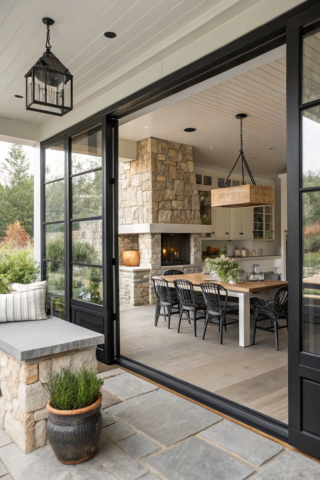 Open black metal-framed sliding glass doors from a stone porch with bench and potted plant into a kitchen dining area with wooden table, chairs, stone fireplace, and white cabinets.