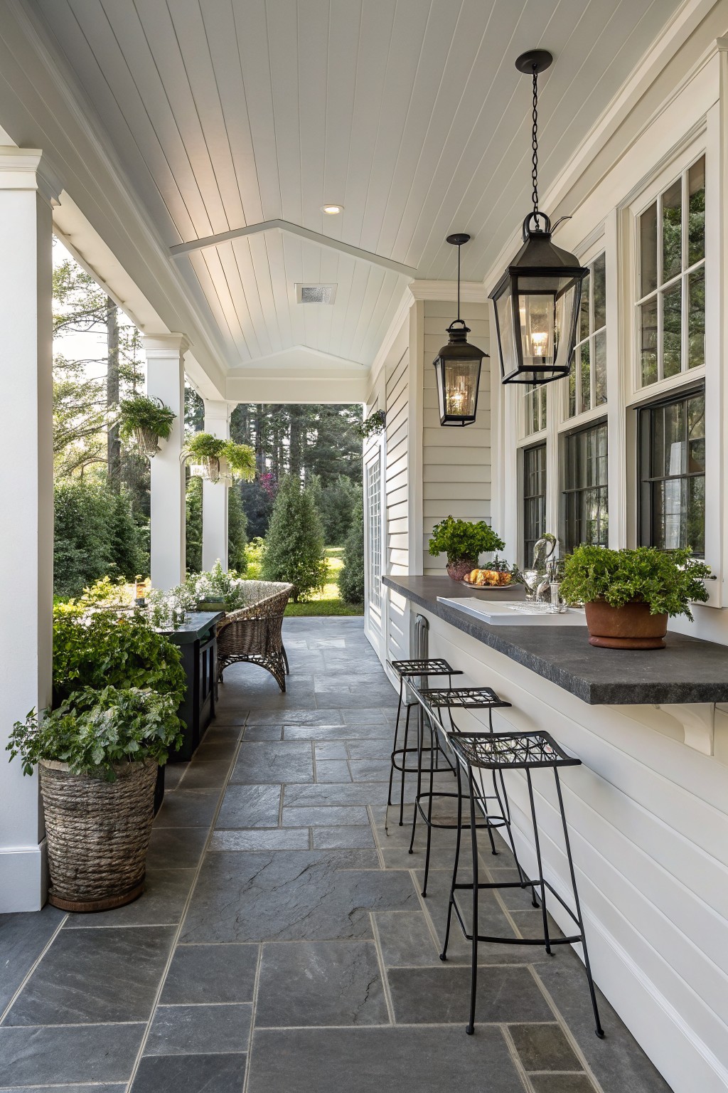 Long covered back porch with white shiplap walls and ceiling, dark countertop bar built against one wall with three black metal barstools, potted plants and glassware on the counter, hanging black lanterns, wicker chair and side table nearby, and trees visible through windows and columns.