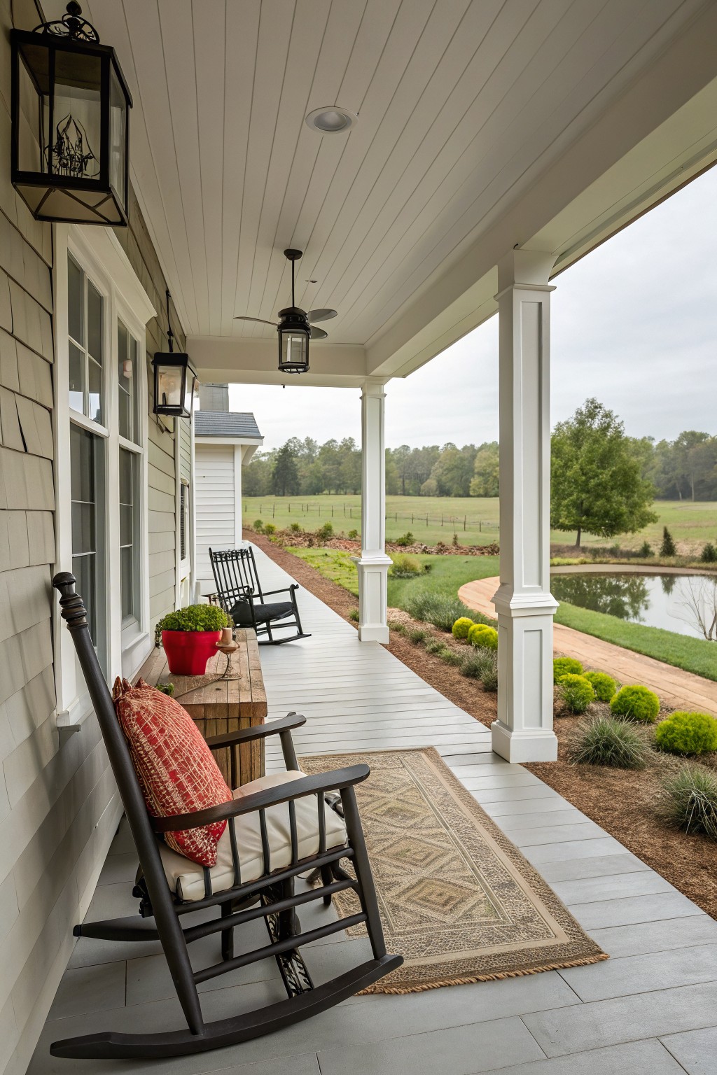 Wide covered back porch on a farmhouse with two black rocking chairs, wooden side table with red potted plant, lanterns, ceiling fan, white columns, overlooking yard, pond, and fields under overcast sky.