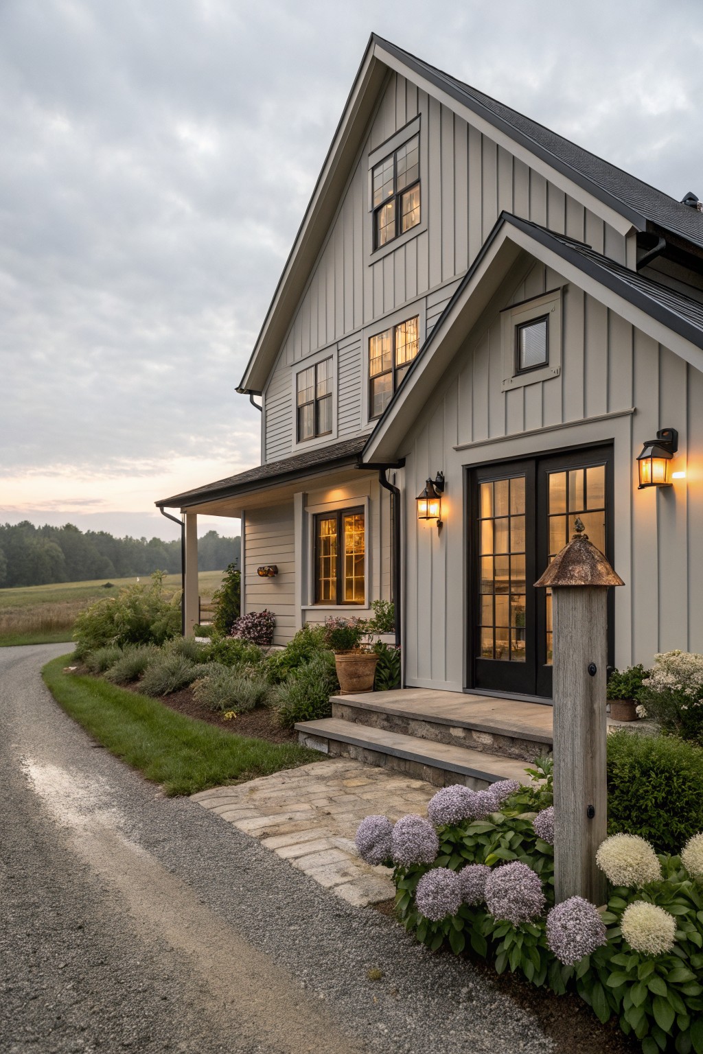 Two-story modern farmhouse exterior with greige board-and-batten siding, gabled roof, black-framed windows and French doors, covered porch, lanterns, stone steps, gravel path, and hydrangea plantings.