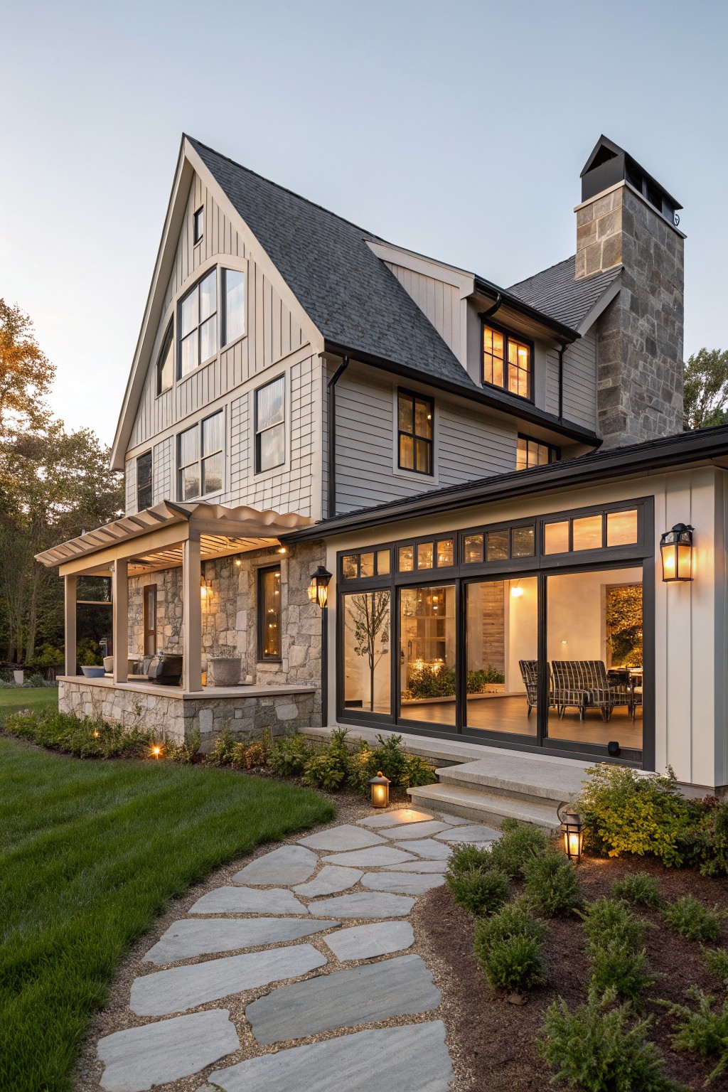 A two-story modern farmhouse exterior featuring greige vertical siding, dark shingled roof, tall gray stone chimney, covered porch with stone base, glass addition, and stone pathway through lawn and shrubs at dusk.