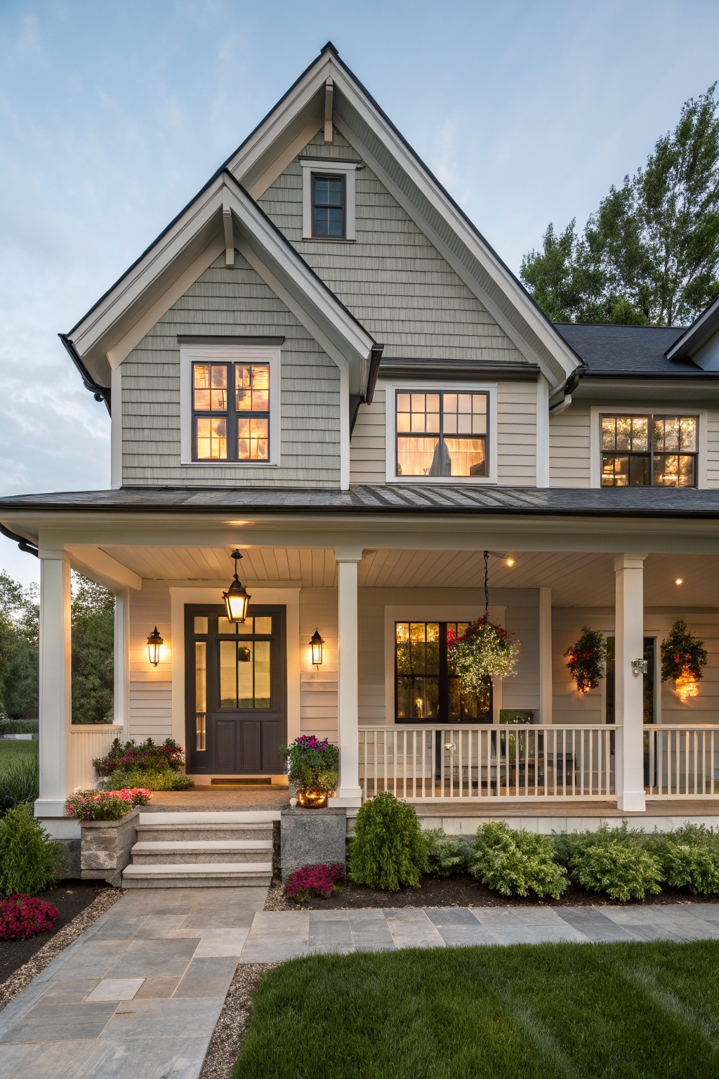 Two-story modern farmhouse exterior with greige shingle siding, white trim, covered front porch, black-framed windows and door, stone steps, and low plantings along a paved walkway at twilight.