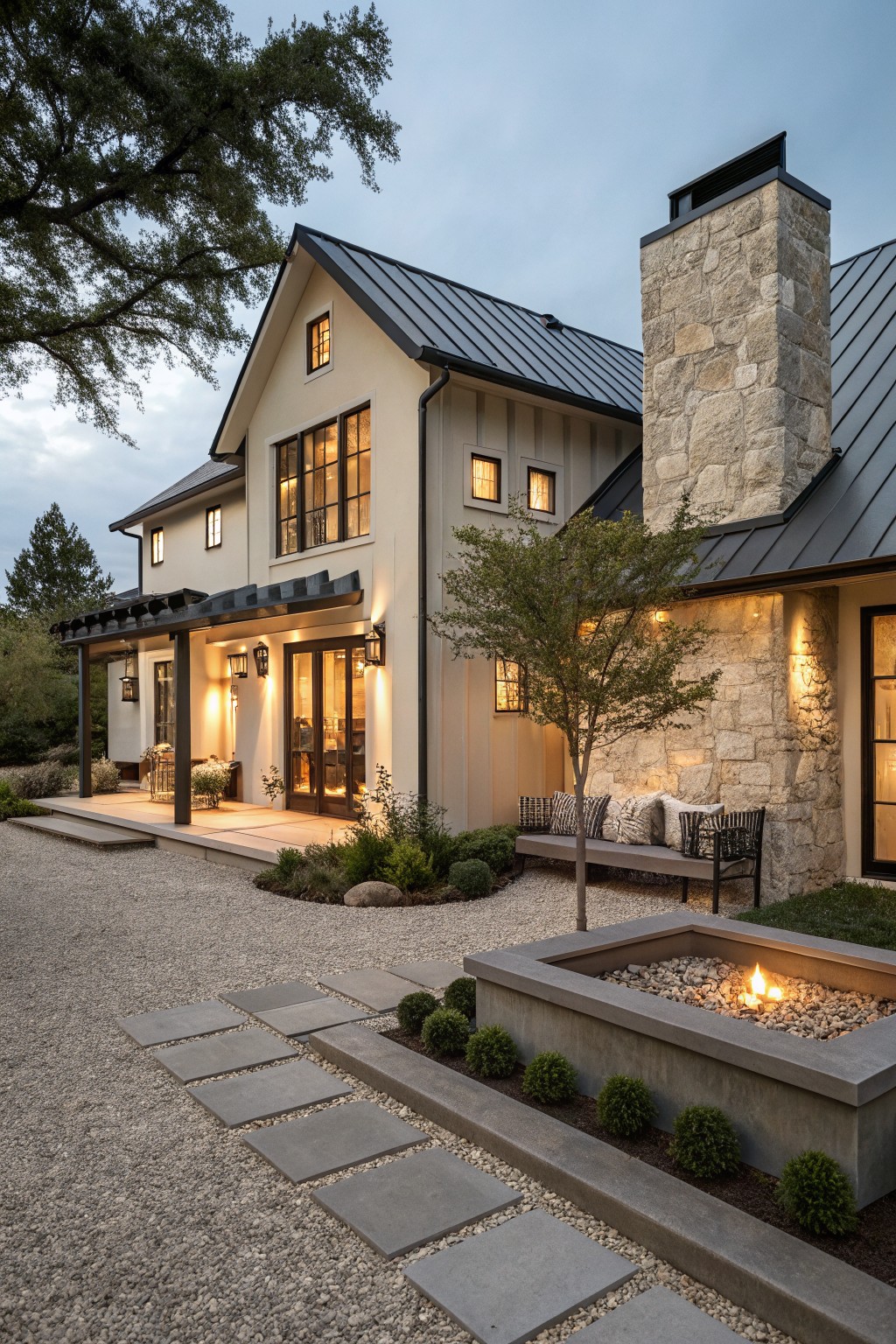 Side exterior view of a two-story greige farmhouse with black metal roof, tall stone chimney, covered porch with glass doors, built-in bench, and linear fire pit in gravel yard surrounded by low plantings.
