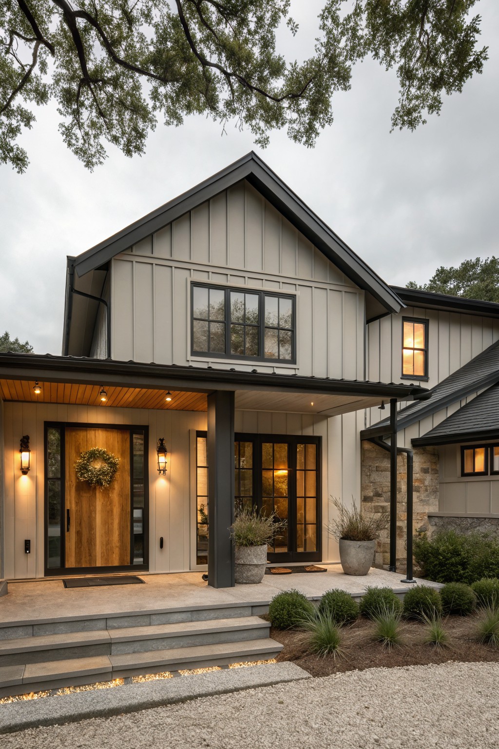 Front exterior of a modern farmhouse house with greige board-and-batten siding, black metal roof and window trim, covered wooden porch, double glass doors, stone side accents, potted plants, and gravel landscaping.