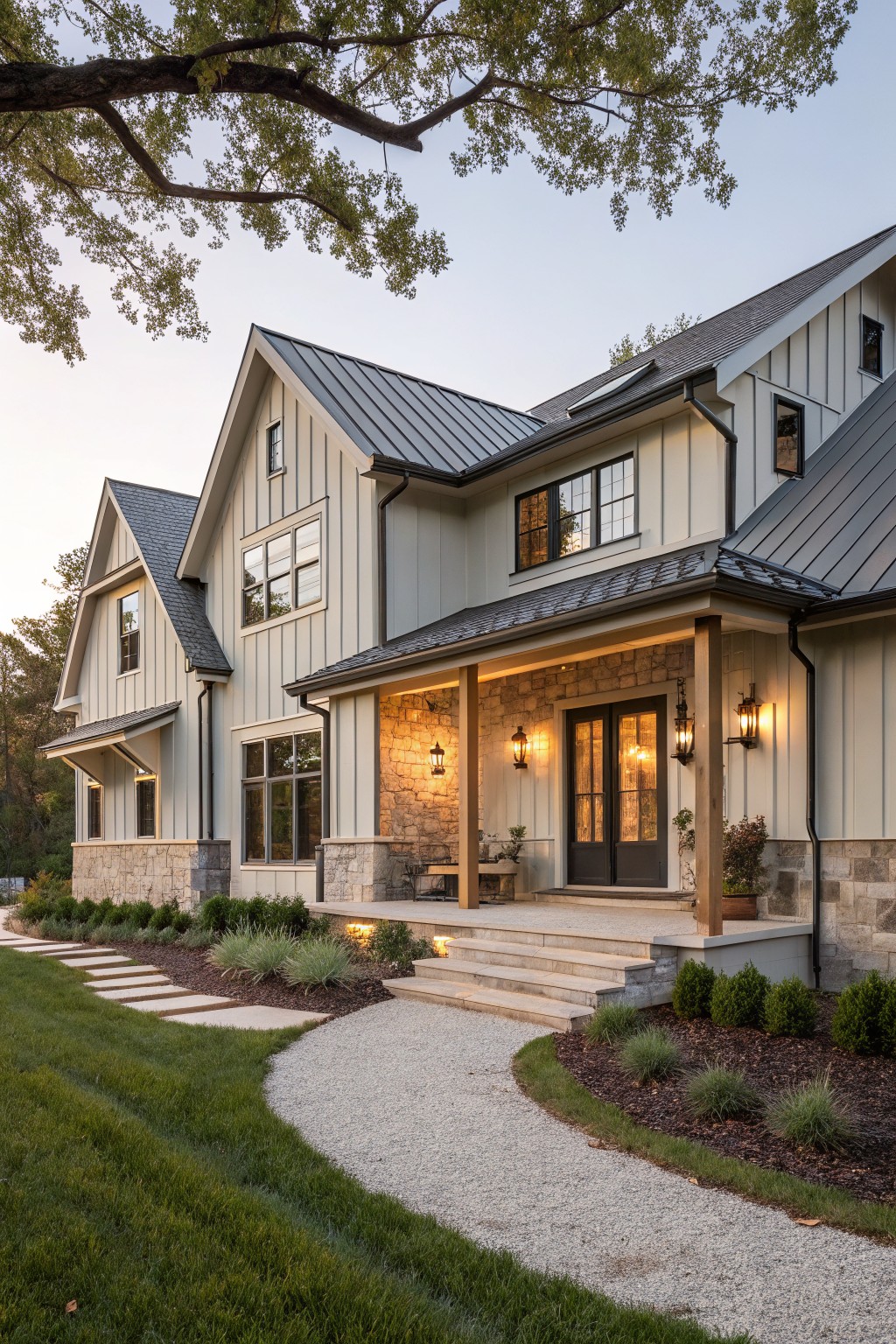 Two-story modern farmhouse exterior with greige board-and-batten siding, dark metal gable roofs, covered front porch supported by stone pillars, large windows, and gravel pathway leading to steps amid landscaped lawn and shrubs at dusk.