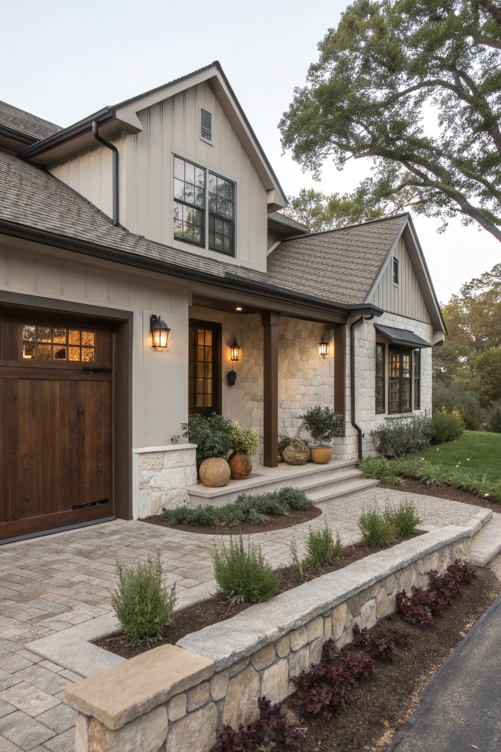Side view of a two-story modern farmhouse with greige board-and-batten siding, wood garage door, stone porch pillars and base, paver walkway, and low plantings along a stone retaining wall.