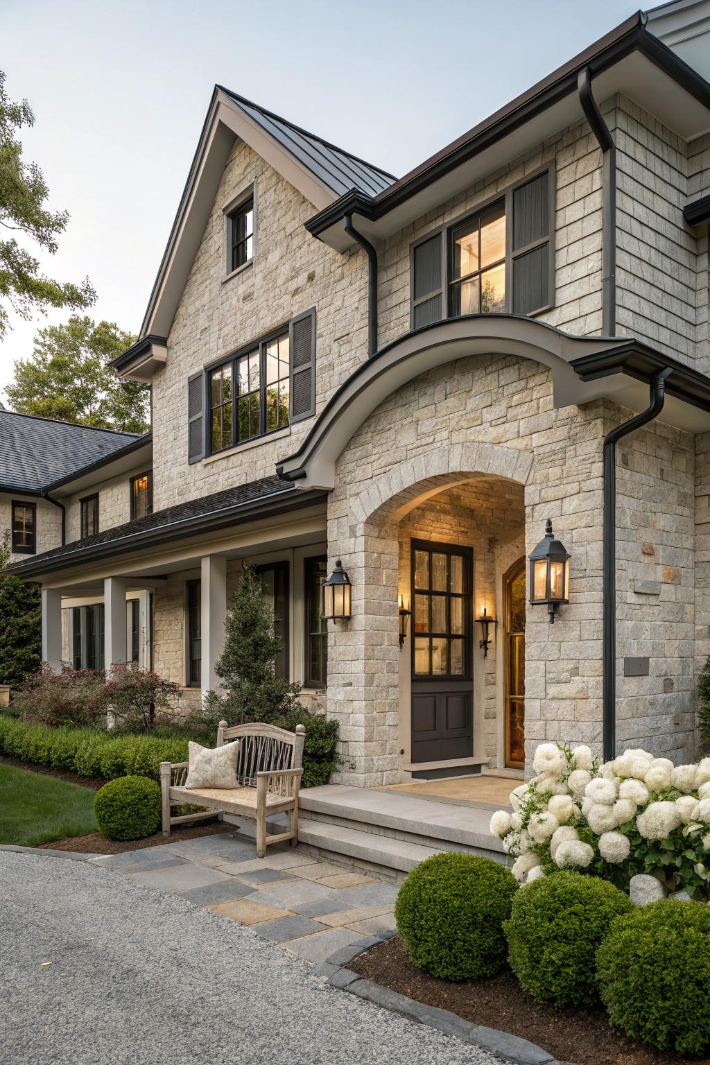 Two-story house exterior with greige stone facade, black-trimmed windows, arched stone entry porch with dark wood door, lanterns, wooden bench with pillow, hydrangeas, boxwoods, and gravel driveway.