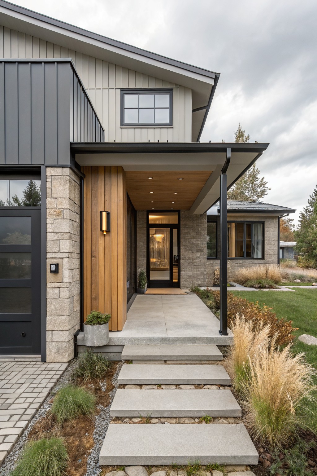 Front exterior of a modern farmhouse house showing a covered entry porch with vertical wood cladding on a post, glass front door, stone corner accents, black metal siding, concrete steps, and ornamental grasses nearby.
