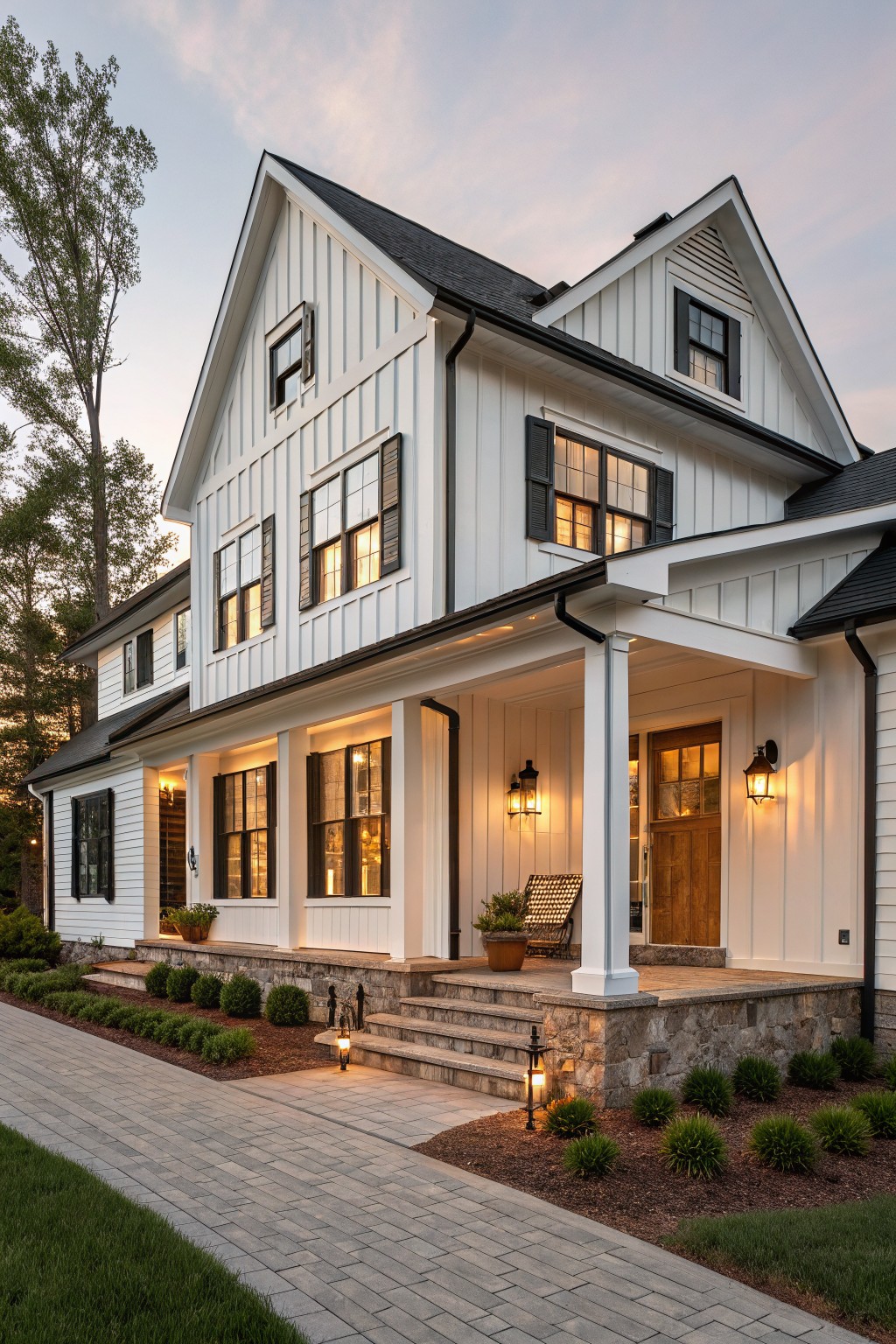 Two-story white board-and-batten farmhouse exterior with black shutters, black roof, wrap-around porch, stone entry steps, lanterns, and low shrubs at dusk.