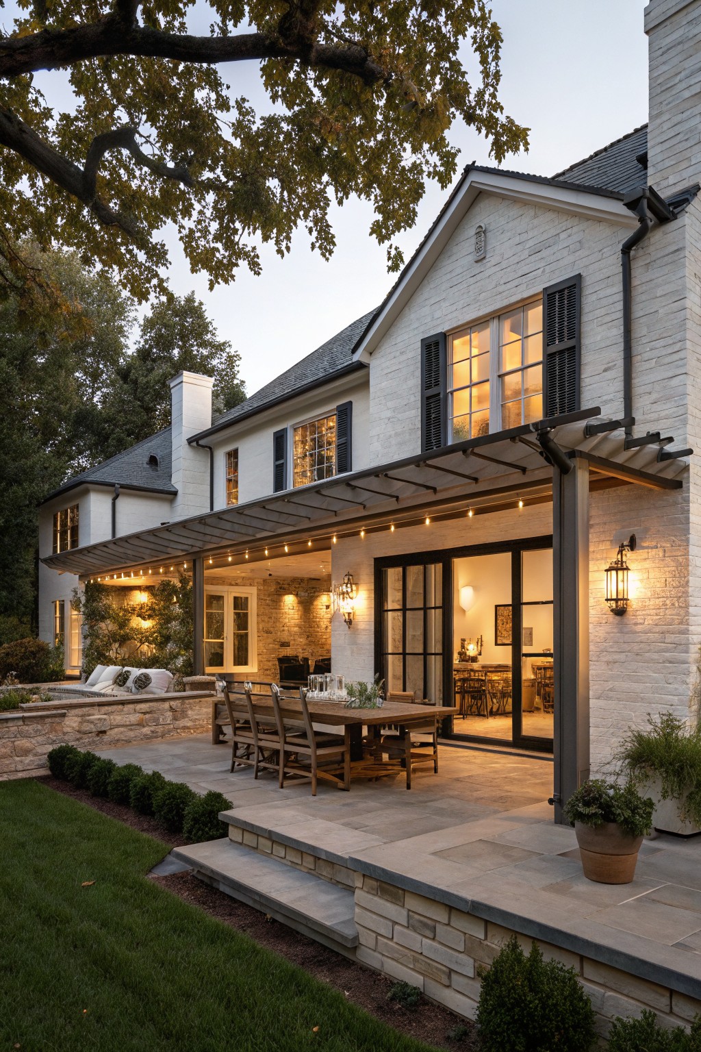 Rear exterior of a two-story white brick house with black shutters on windows, a metal-roofed covered patio with string lights and wooden dining table, stone steps, and landscaped yard.