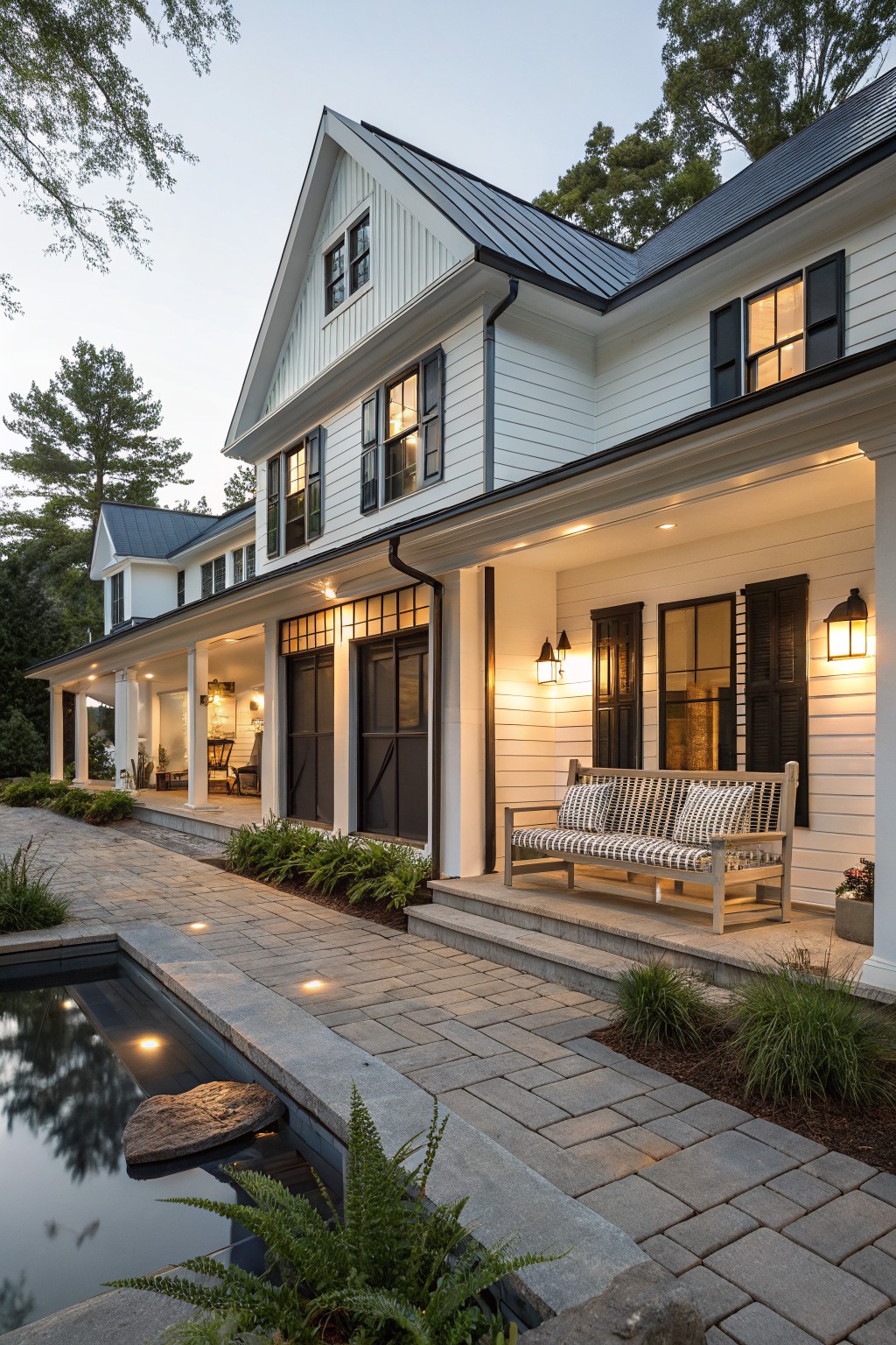 White modern farmhouse exterior with black shutters, wraparound porch, metal roof, pathway and small pool in the foreground, trees and dusk lighting.