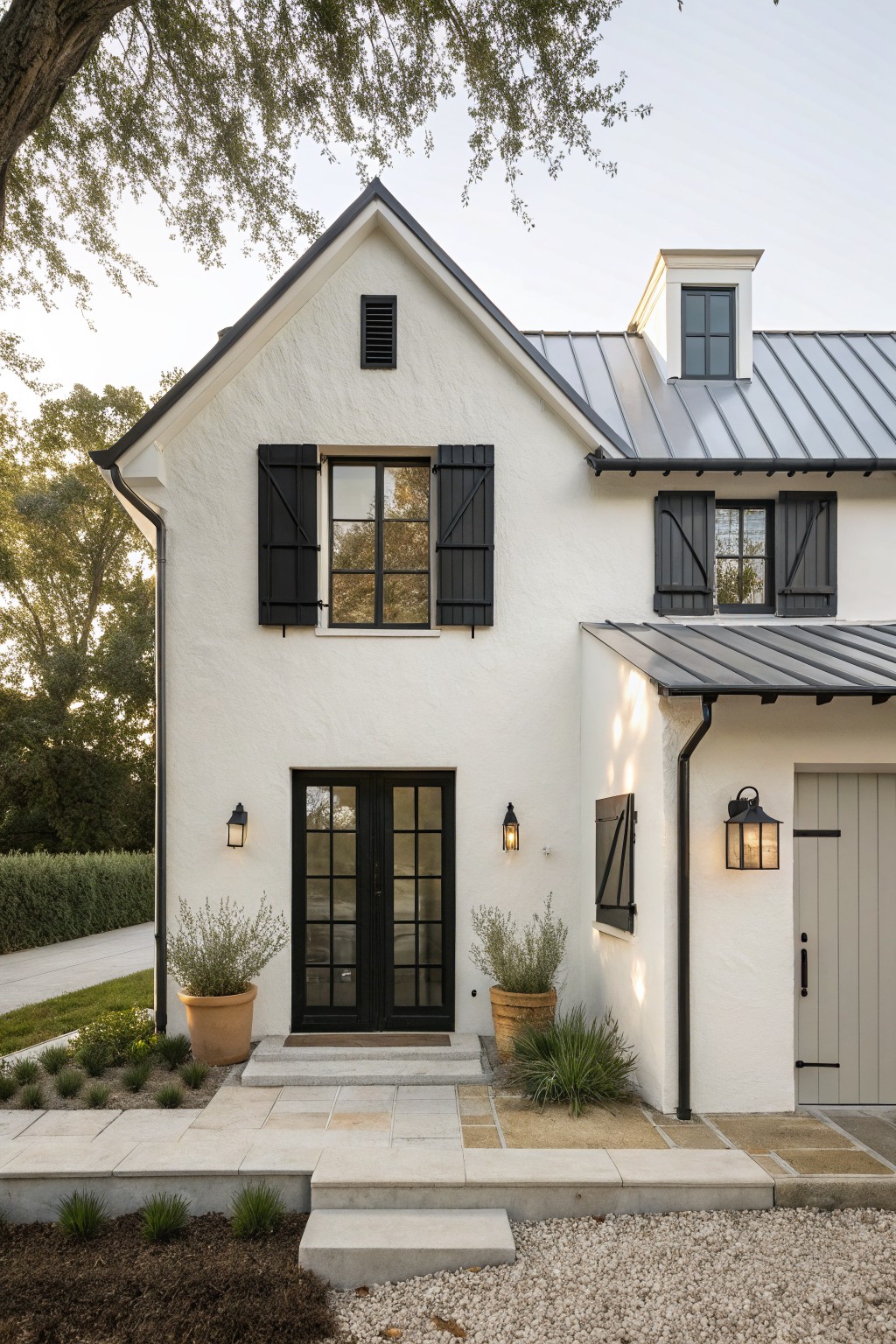White stucco modern farmhouse exterior with black board-and-batten shutters on multi-pane windows and French doors, gray standing-seam metal roof, lantern wall lights, potted plants flanking the entry, stone steps, and gravel path.