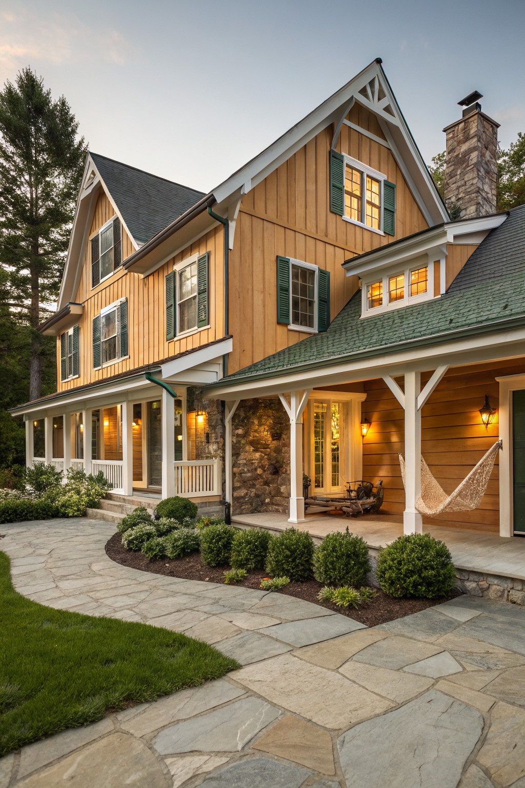 Two-story modern farmhouse exterior with golden board-and-batten cedar siding, green louvered shutters on white-trimmed windows, wrap-around porch with white posts and hanging lantern lights, stone foundation and chimney, hammock on porch, flagstone path, shrubs, and tall pine trees at dusk.