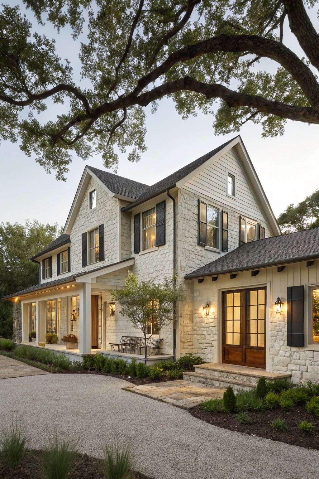 Two-story modern farmhouse exterior with white stone base, light clapboard siding, black shutters on multipaned windows, covered front porch with columns, wooden double doors, lanterns, gravel driveway, low plantings, and large overhanging oak tree branches at dusk.