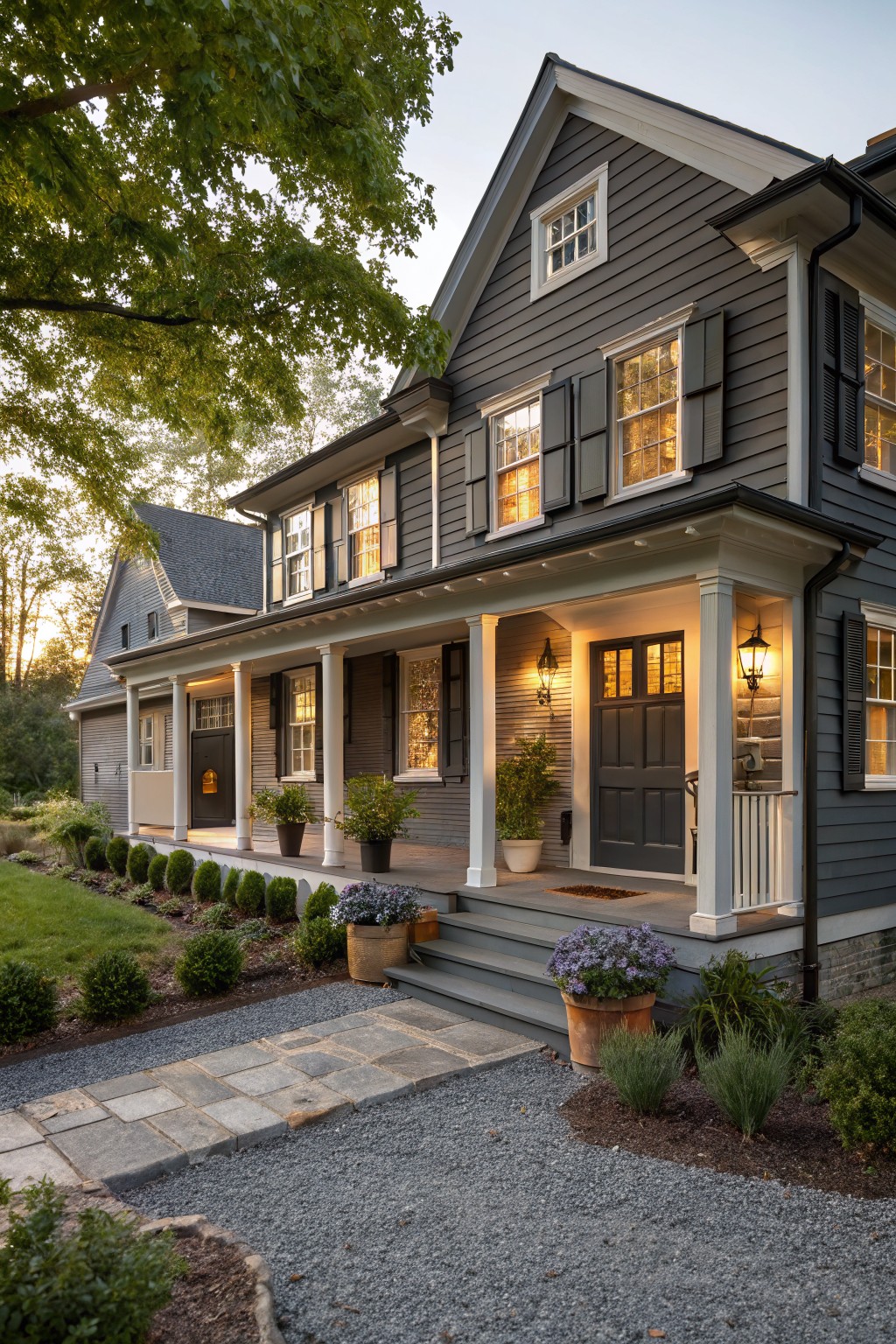 Two-story gray shingled house exterior with black shutters on windows, white-trimmed porch supported by columns, lanterns flanking a dark wood entry door, potted plants on the porch steps, stone pathway, and low landscaping in evening light.