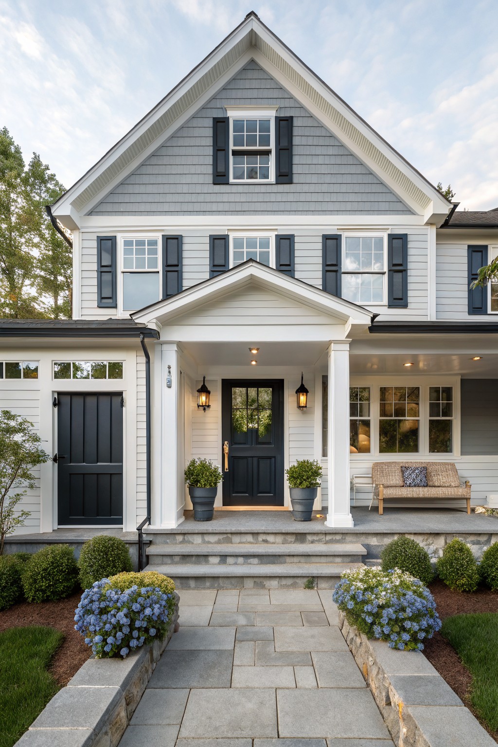 Front view of a two-story light gray shingled house with black shutters on white-trimmed windows, a white gabled portico with black front door, stone steps leading to the entry, potted plants on the porch, and blue flowering shrubs along a flagstone path bordered by stone walls.