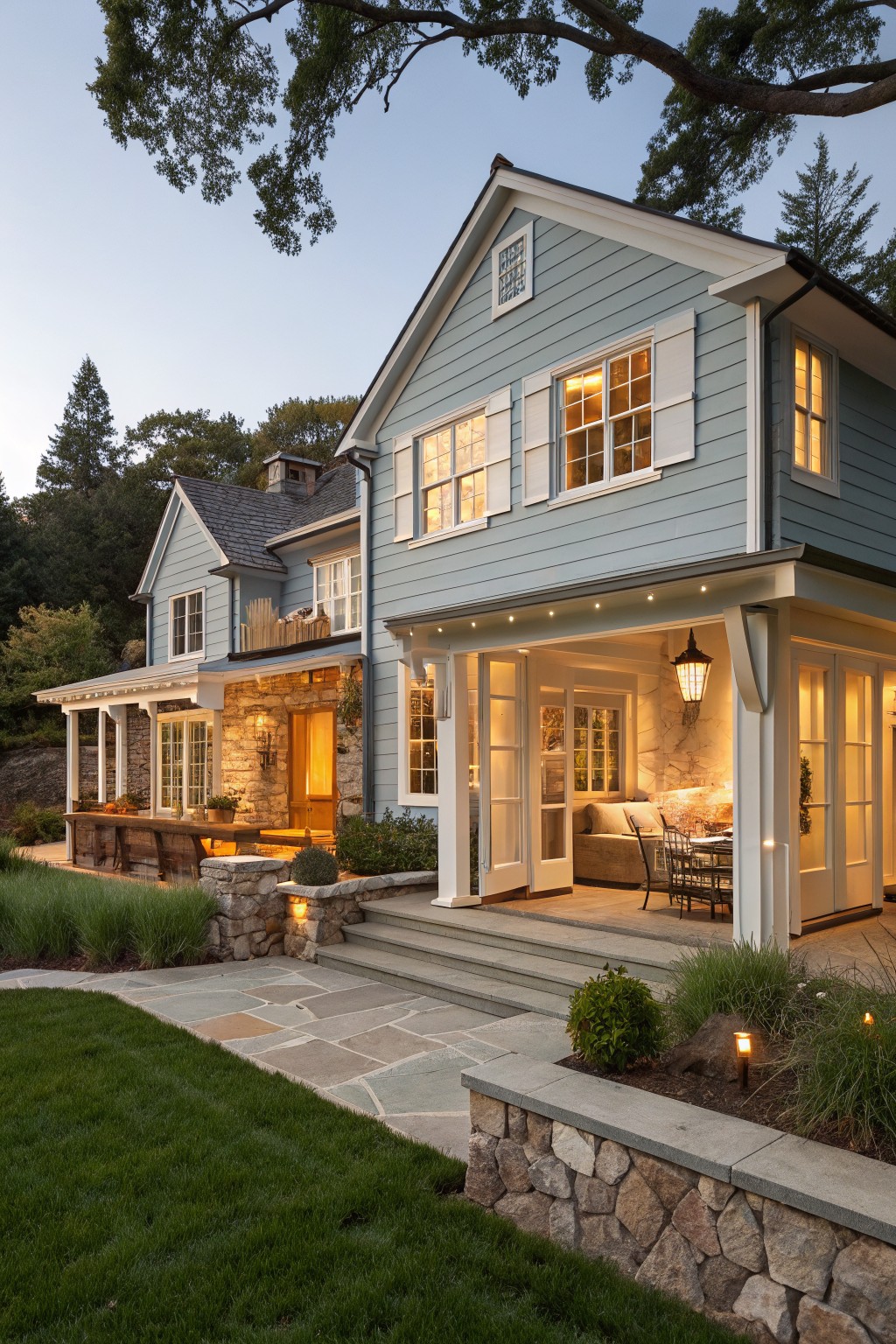 Light blue clapboard two-story house with white shutters and trim, stone-based wooden porch, open patio with French doors and furniture, pathway, low stone wall, and landscaping at evening.