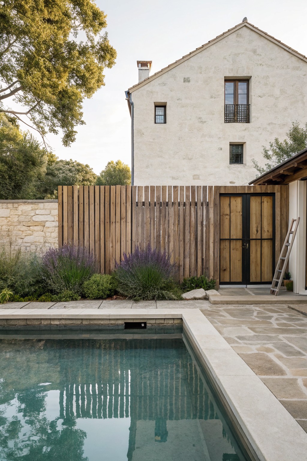 Turquoise pool with beige stone coping and edge, next to tall vertical slatted wooden fence, lavender plants, stone wall, and white stucco house with wooden gate.