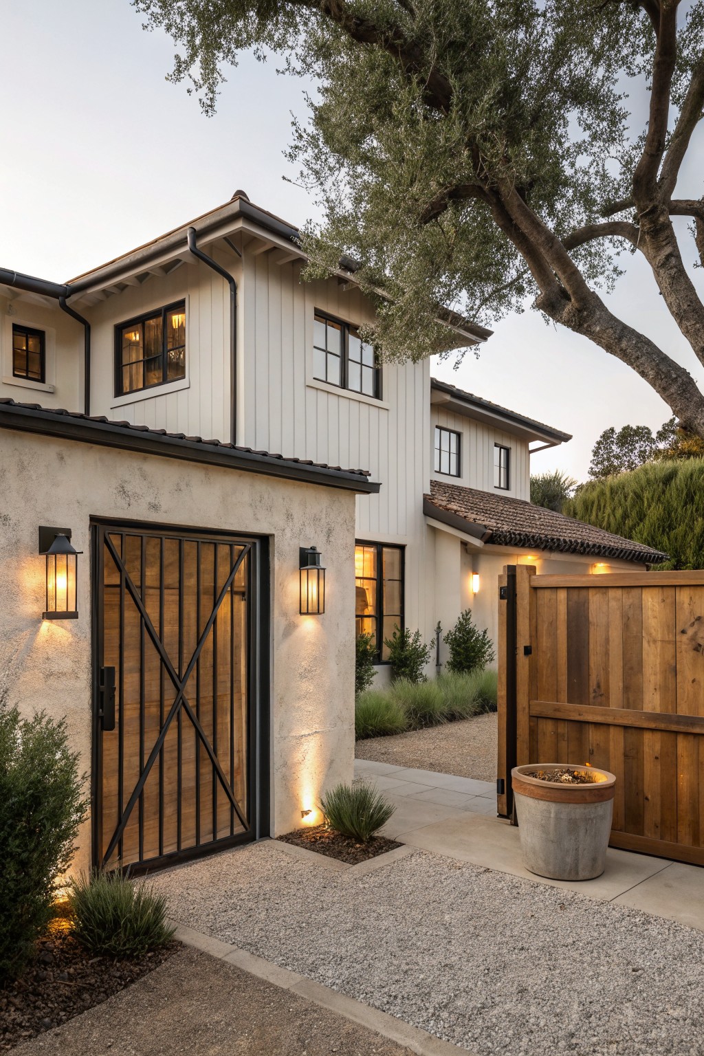 Exterior view of a modern farmhouse home showing a black metal bar gate with crossbars on a stucco wall next to a closed vertical slat wooden fence gate, gravel pathway, potted plant with fire, low shrubs, and wall-mounted lanterns under an olive tree.