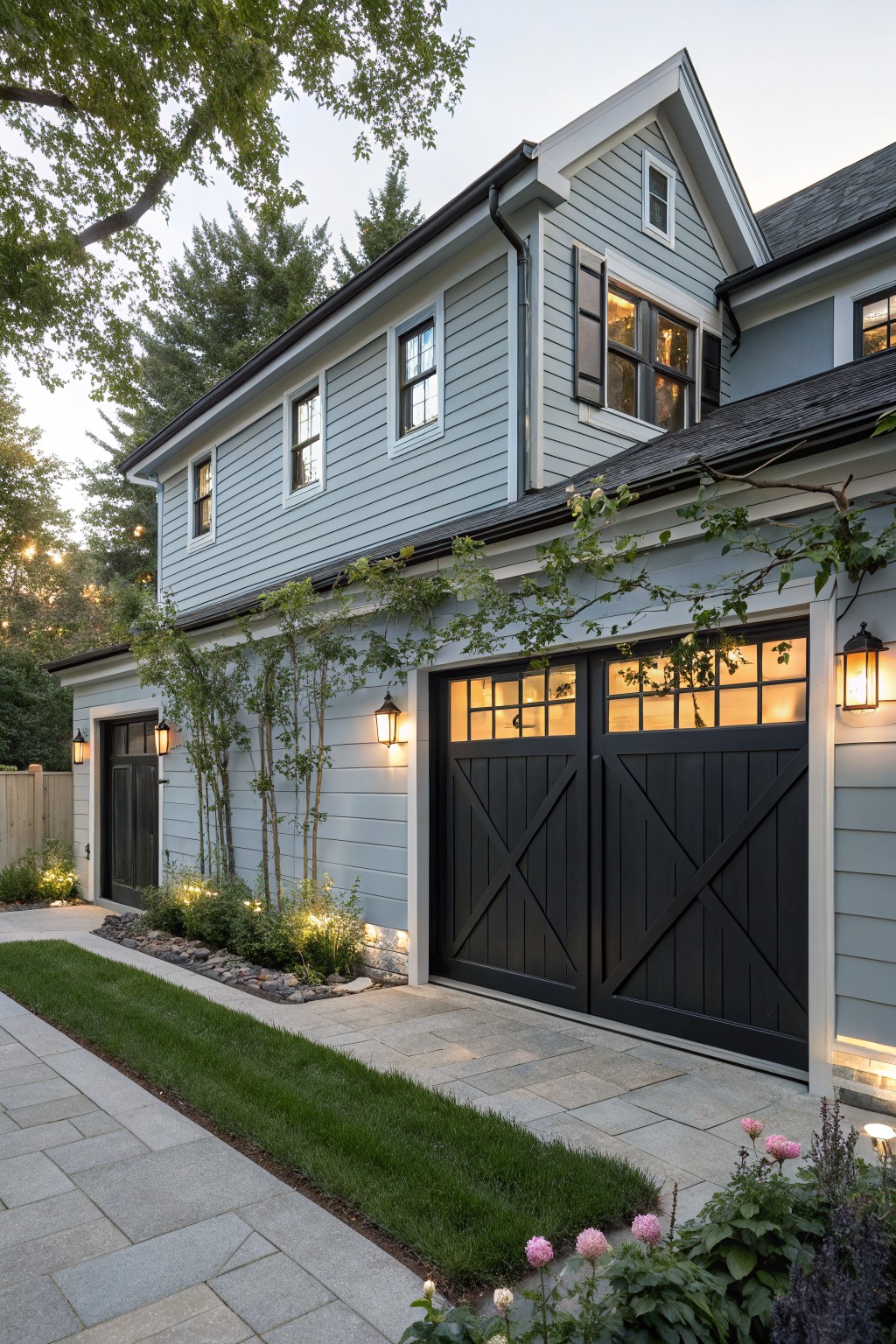Side exterior view of a two-car garage on a light blue clapboard house with black double doors, flanked by black lantern wall sconces, young trees and plants nearby, stone pathway, and landscape lighting.