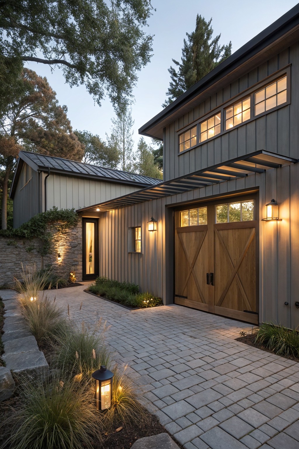 Exterior of a two-car garage with wooden paneled doors, gray board-and-batten siding, black lantern wall lights on either side of the doors, a nearby side entry door, paver stone pathway edged with grasses and stone lanterns, surrounded by trees at dusk.