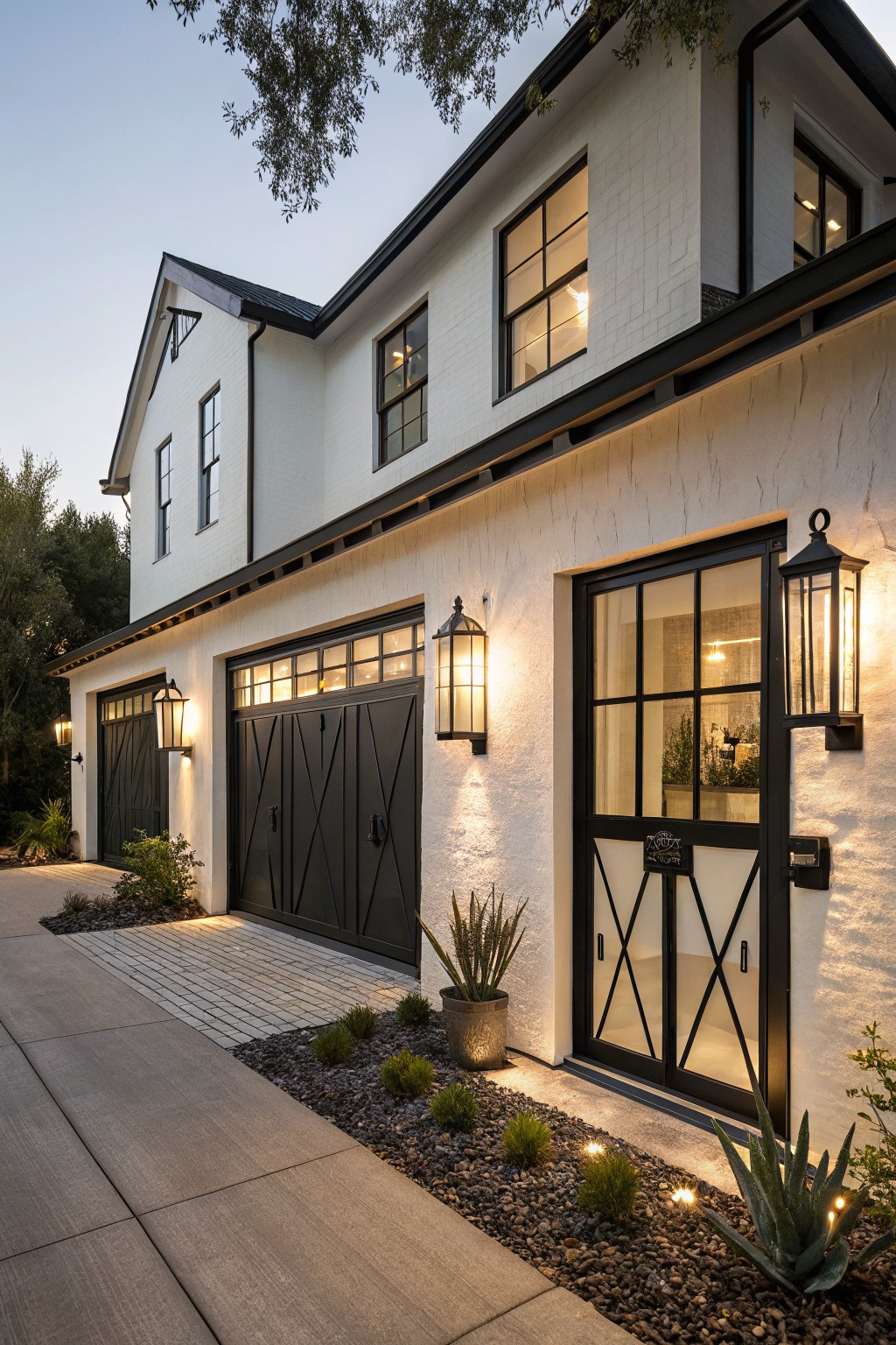 White stucco modern farmhouse house exterior at dusk featuring black X-pattern garage doors, a glass-paneled entry door, large black lantern lights, and gravel landscaping along the driveway.