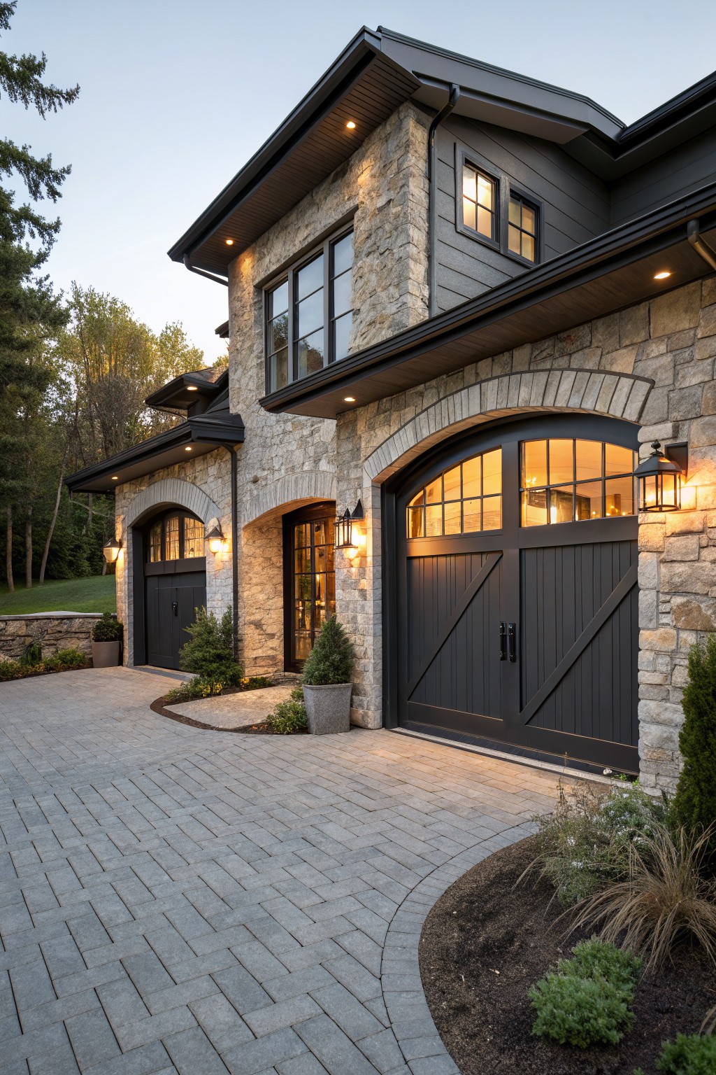 House exterior with two arched black garage doors on stone walls, flanked by black lantern wall lights, plus a curved paver driveway and low plants at dusk.