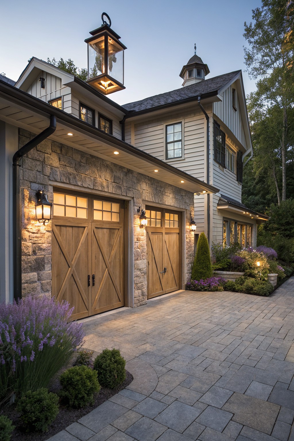 Side view of a modern farmhouse garage featuring two wooden X-braced doors flanked by black lantern lights on stone walls, with recessed soffit lights, purple lavender plants, boxwoods, and a gray paver driveway at twilight.