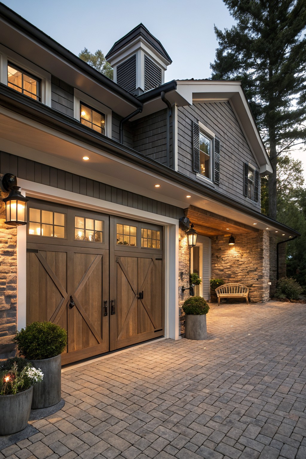 Side exterior of a modern farmhouse house featuring a two-car garage with dark wood X-brace doors, black lanterns on stone pillars, paver walkway, potted plants, and warm lighting at dusk.