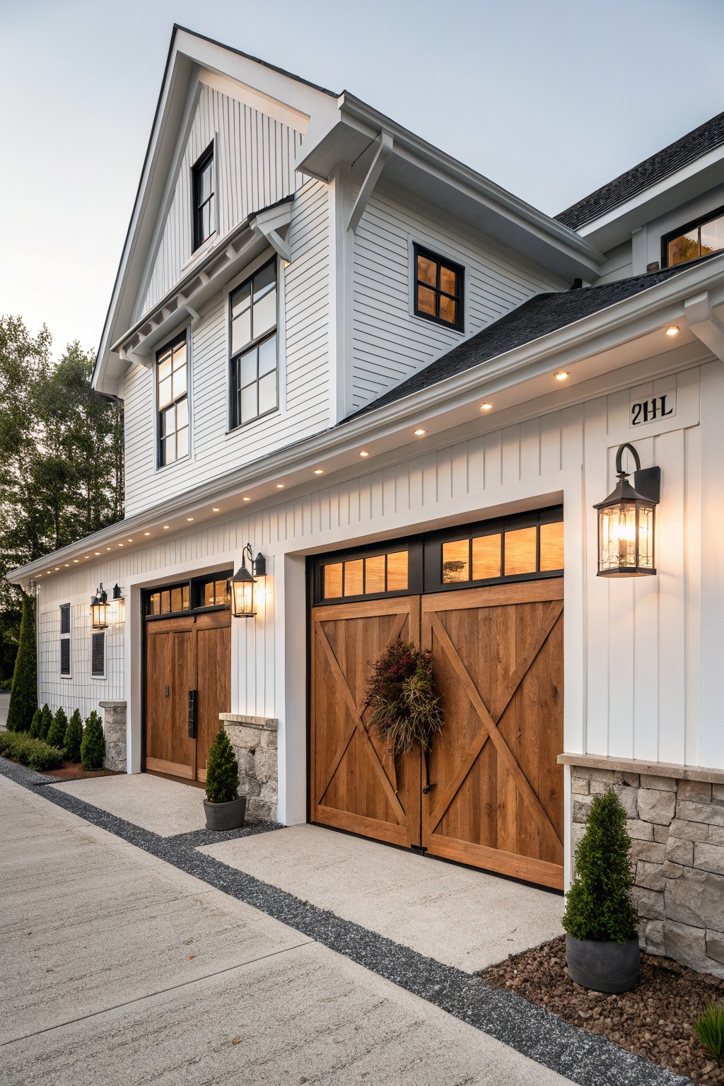 White shiplap-sided modern farmhouse house exterior at dusk with double wooden garage doors, black-framed windows, wall-mounted black lantern lights, recessed eave lights, stone accents, potted plants, and a concrete driveway edged in gravel.