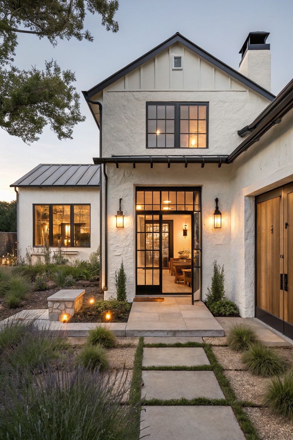 One-story modern farmhouse house exterior at dusk with white stucco walls, black metal-framed glass entry doors and windows, wooden garage door, metal-roofed garage wing, lantern lights, and gravel pathway with grasses and stone bench.