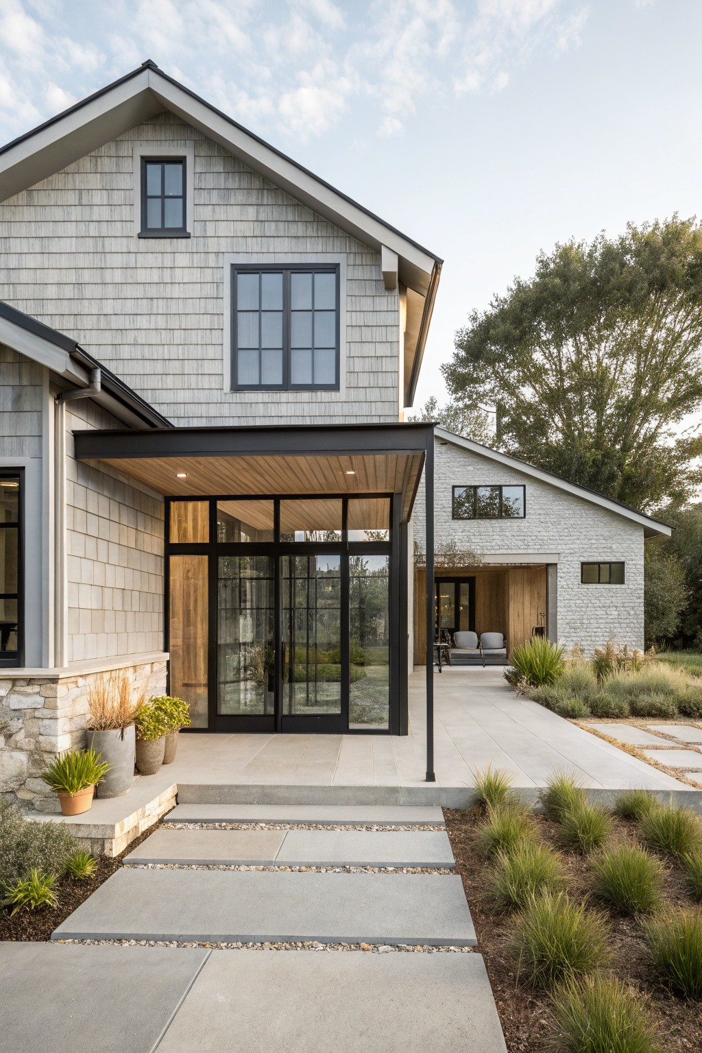 One-story modern farmhouse exterior with light gray shingle siding, black steel canopy over wood-ceilinged entry with glass doors, stone accents, concrete pathway, and grasses.