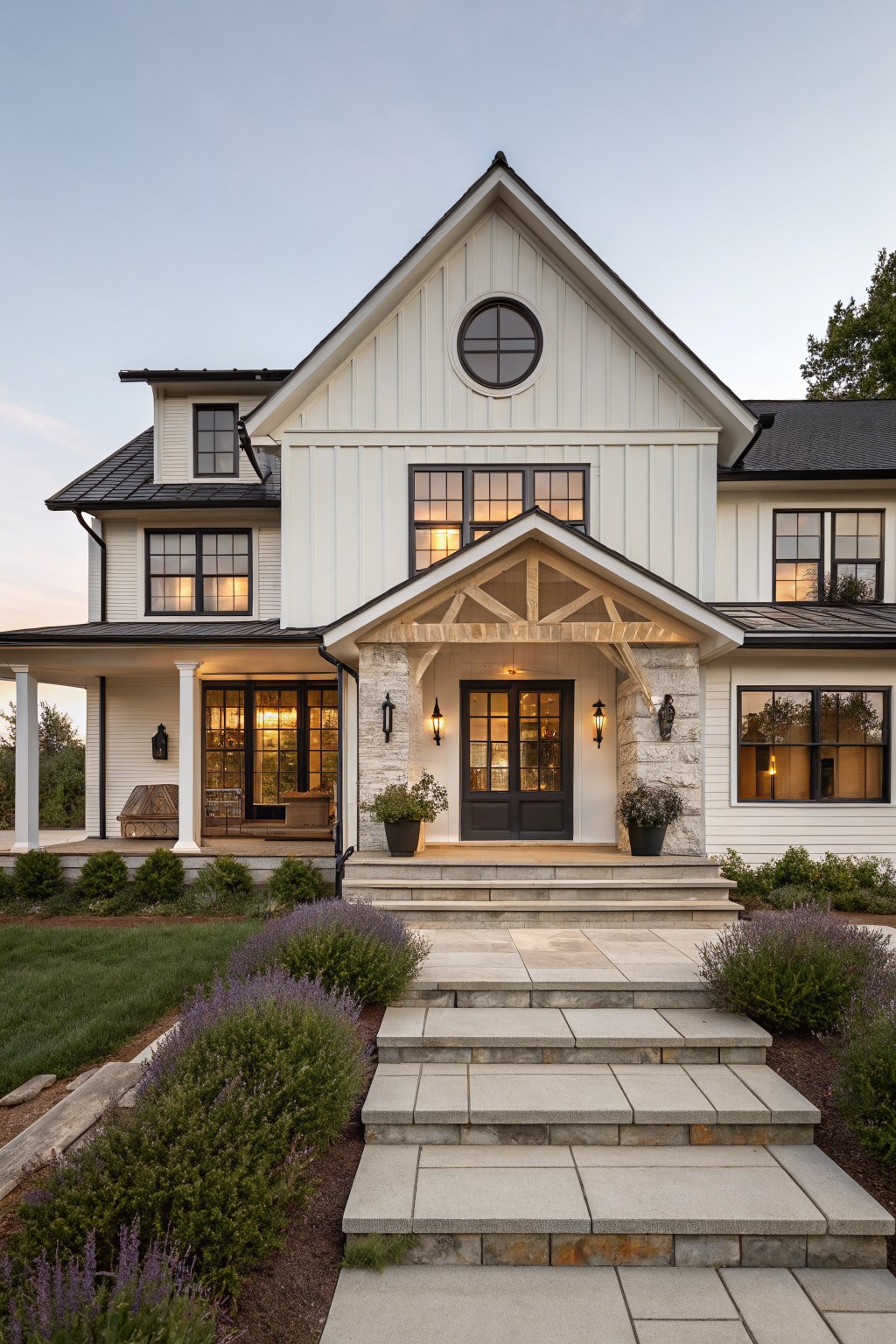 Front view of a two-story modern farmhouse house with white board and batten siding, black metal roof, black-framed windows, a gabled entry porch with wooden truss beams and stone pillars, black front door, porch lighting, and stone steps flanked by lavender shrubs.