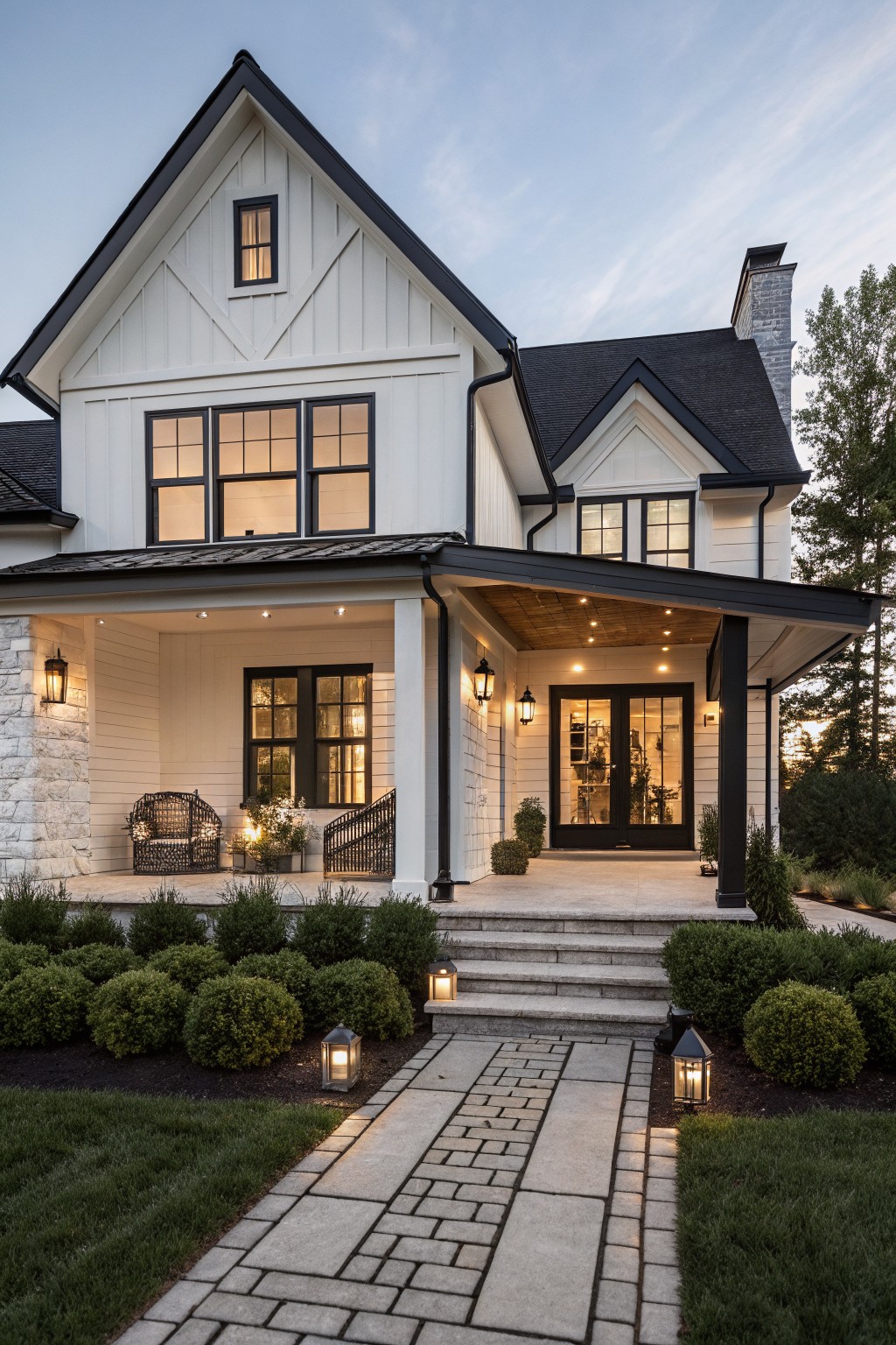 One-story modern farmhouse exterior featuring white board-and-batten siding, black gabled roof and trim, large black-framed windows, covered porch with stone pillars and lanterns, stone steps, brick pathway lined with lanterns and boxwood shrubs, and lawn at dusk.