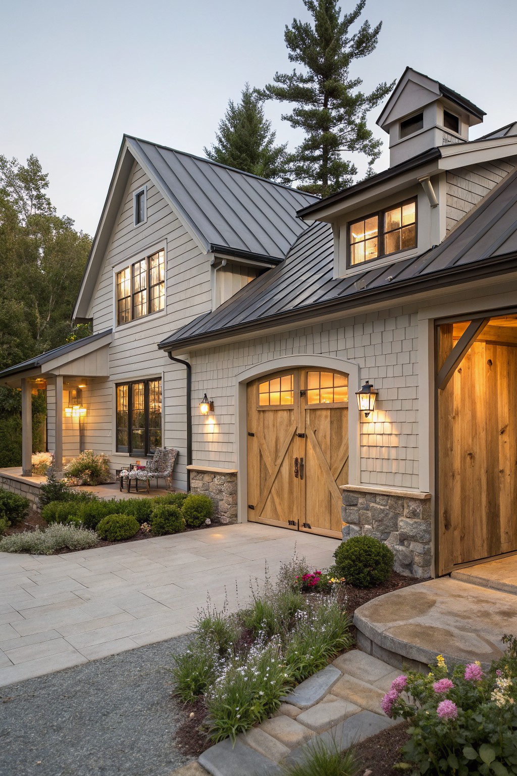 Exterior of a one-story modern farmhouse with light gray board-and-batten siding, dark standing-seam metal roof, large wooden garage doors with arched glass panels, adjacent porch, stone foundation, lanterns, and gravel driveway edged by plants at twilight.
