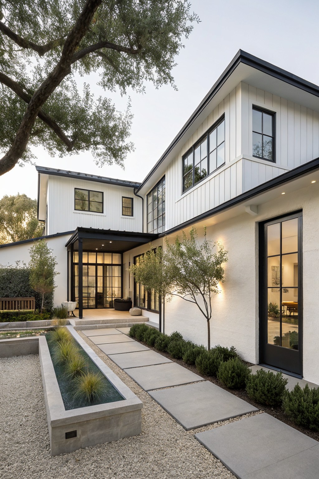 A two-story modern farmhouse house with white vertical board-and-batten siding, black trim on large windows and doors, a covered entry porch with black metal framing on glass doors, and a concrete walkway edged by grasses and shrubs in gravel.