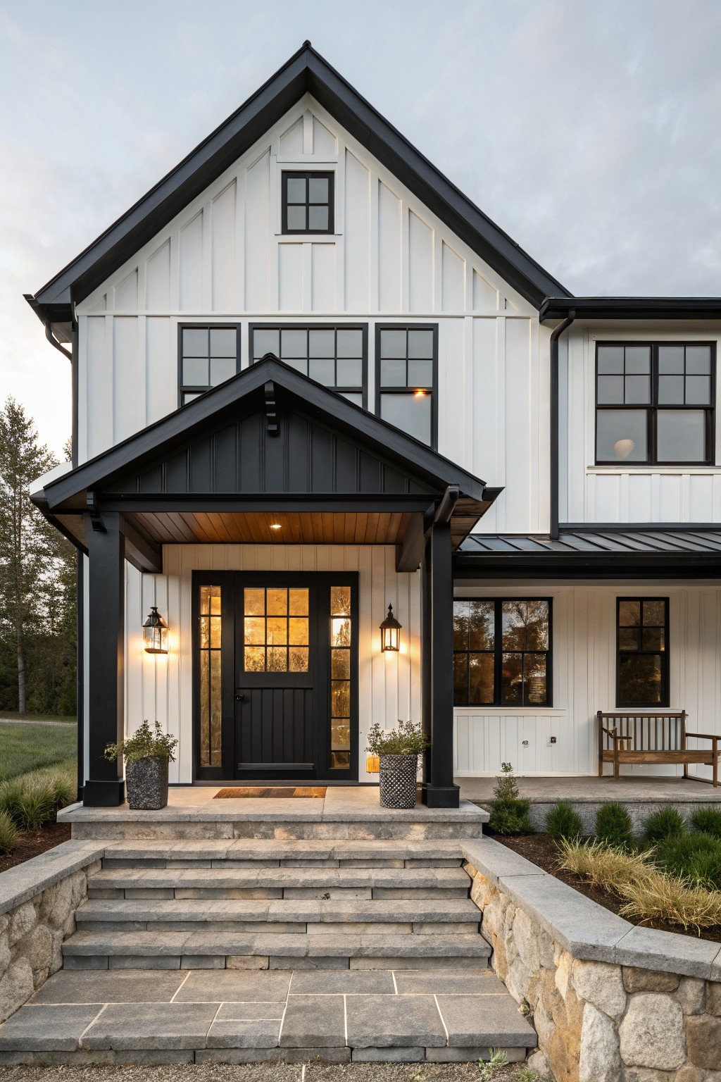 Front view of a modern farmhouse house with white board-and-batten siding, black trim on windows and gables, covered gabled porch with black door and lanterns, stone steps, retaining wall, and low plantings.