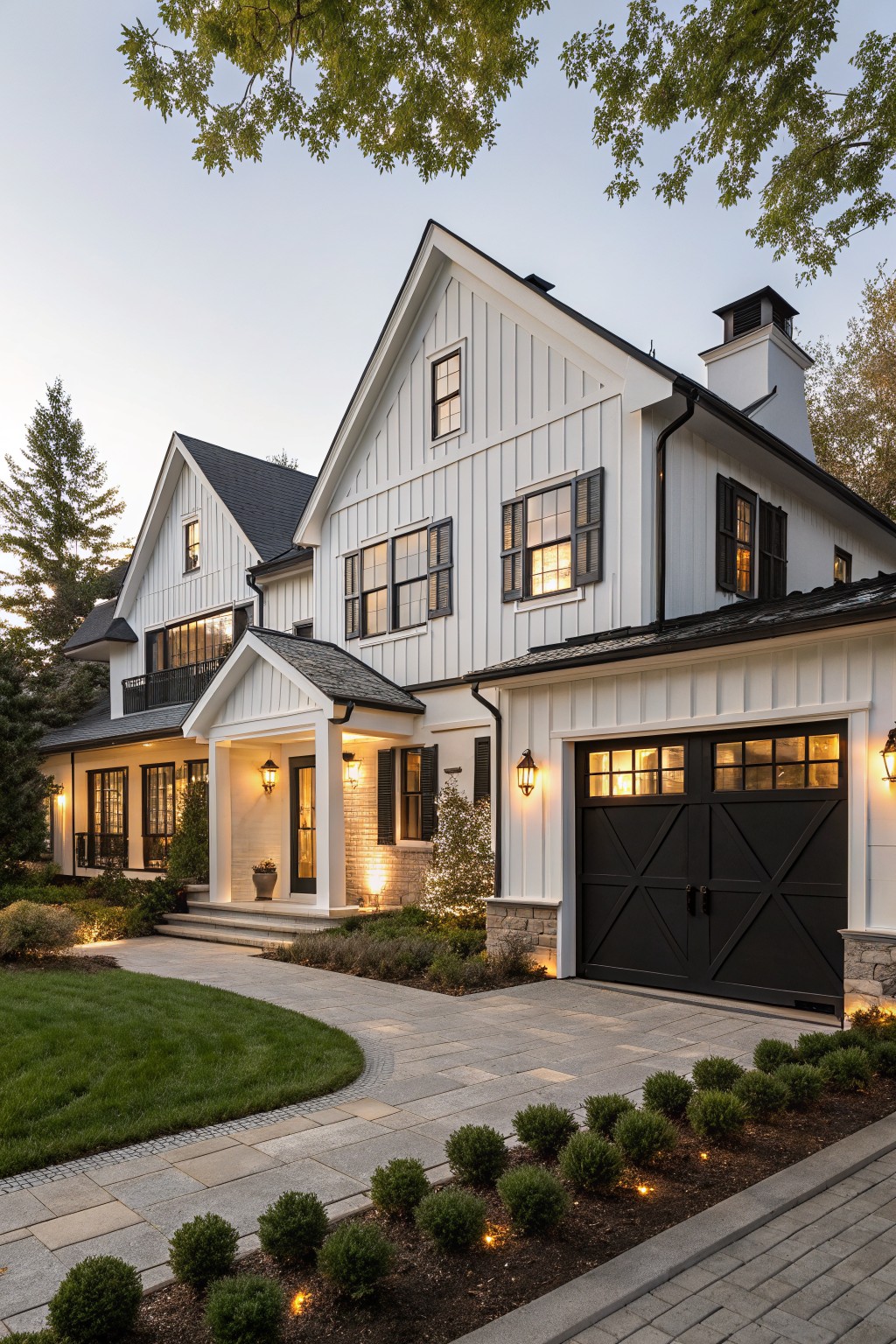 Two-story white board-and-batten house exterior with black window trim and shutters, black X-pattern double garage doors, covered entry porch with lanterns, stone pathway, boxwood edging, and trees at dusk.